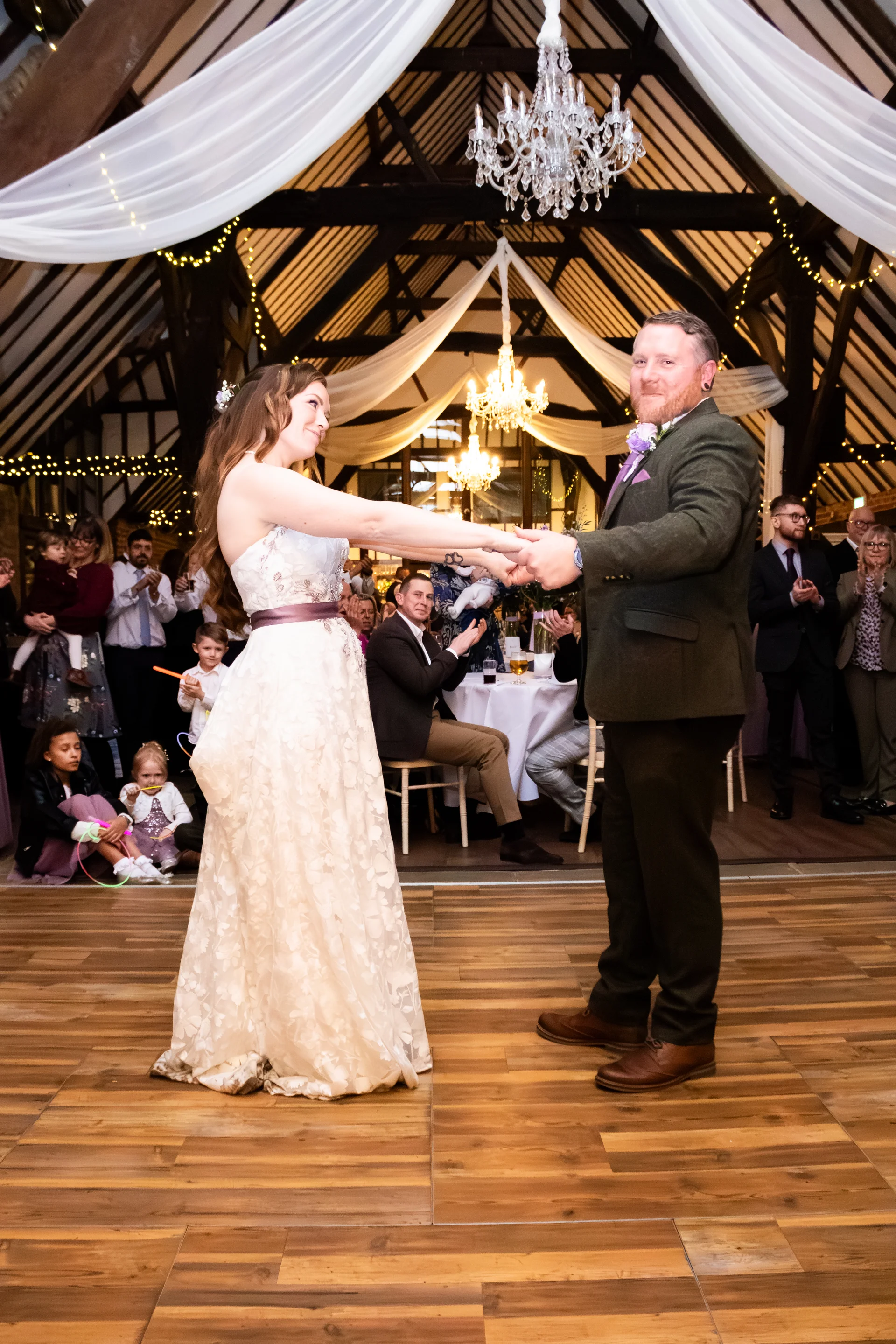 Newlyweds share their first dance on a wooden floor inside a rustic barn venue with exposed timber beams and sparkling fairy lights.