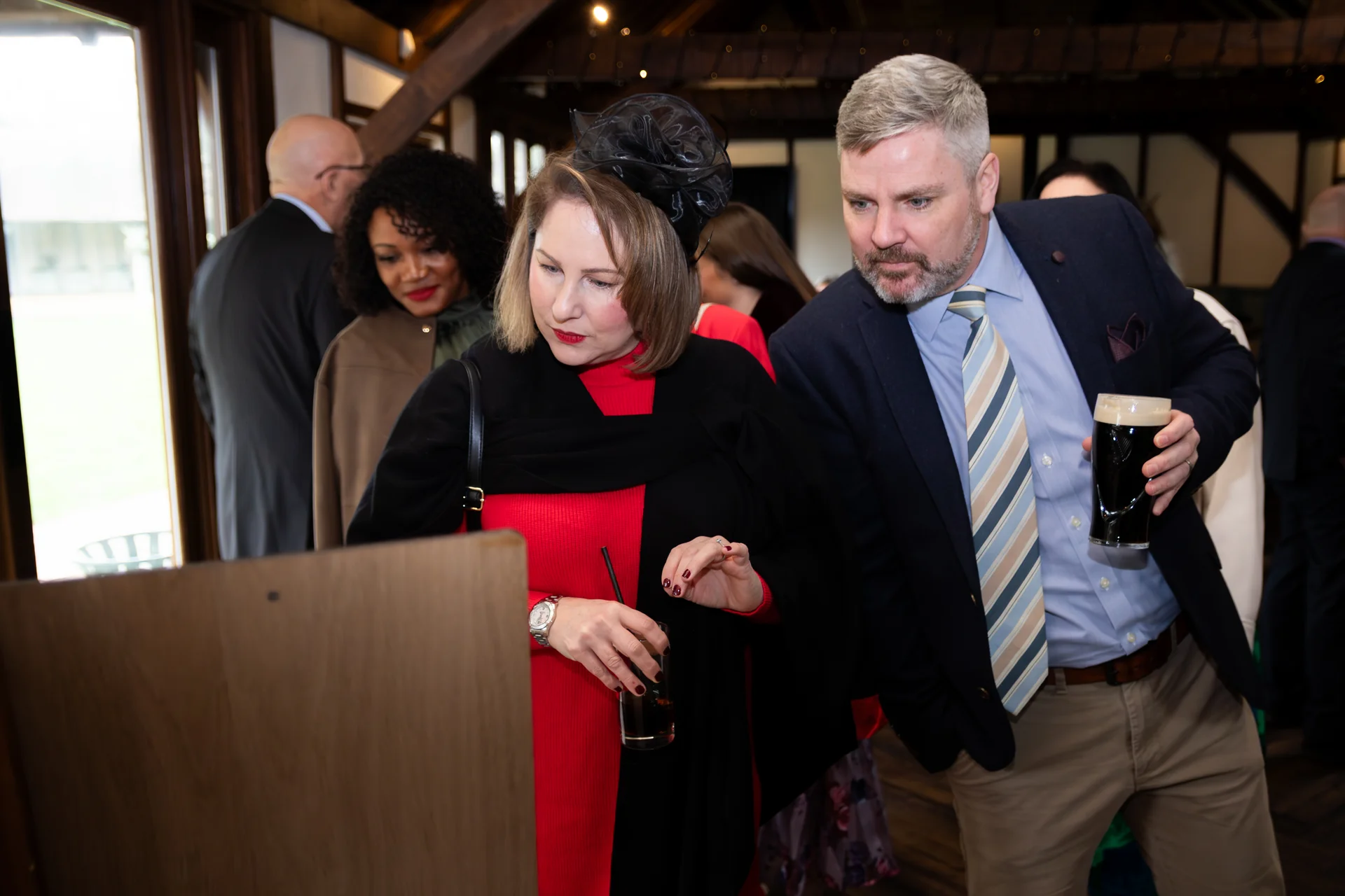 Wedding guests in a rustic barn venue lean in to look at something together, with a woman in a red dress wearing a black fascinator and a man in a blazer holding a pint of stout.
