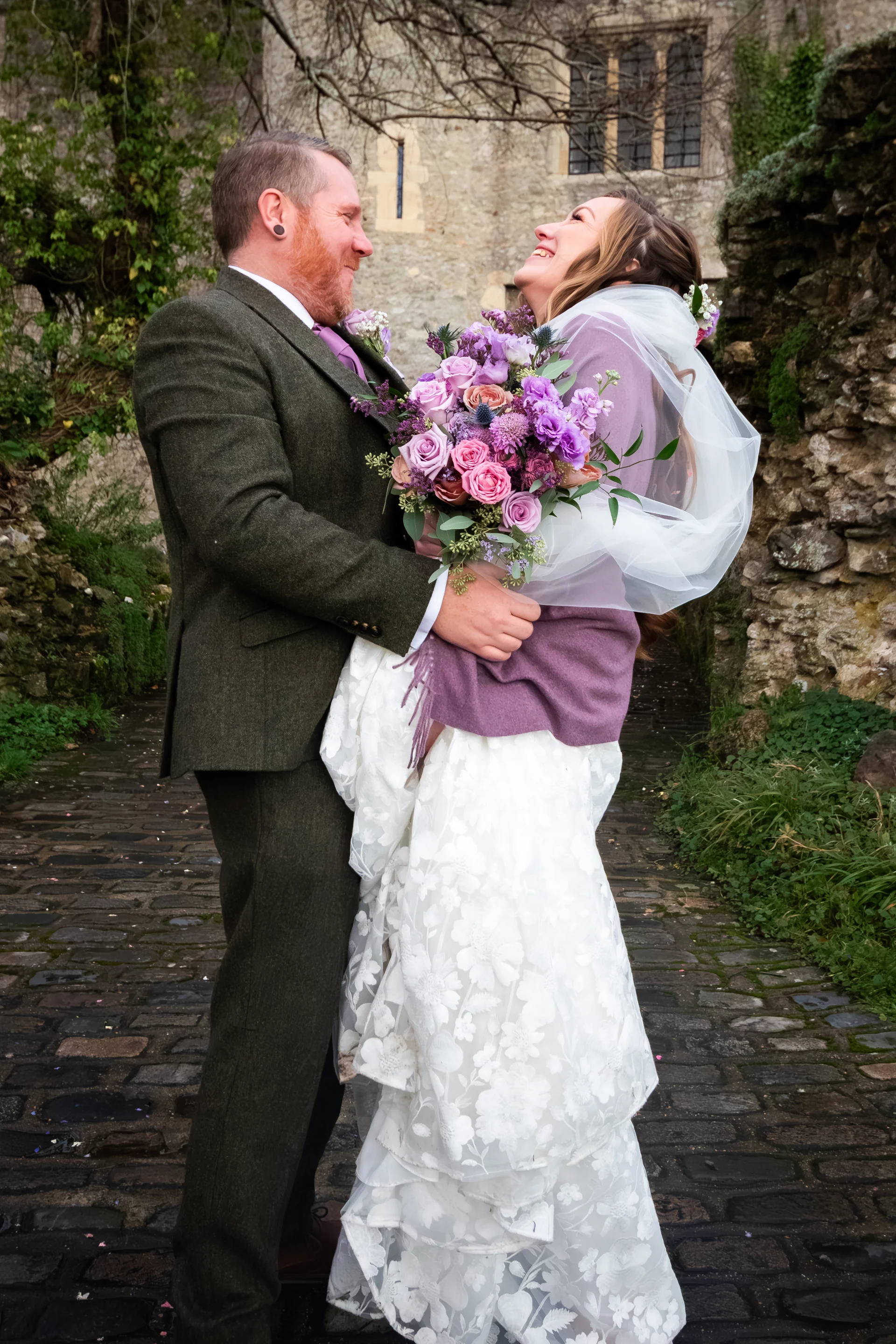A groom in an olive tweed suit happily lifts his bride in the air as they laugh together on a cobbled path outside a historic stone castle.
