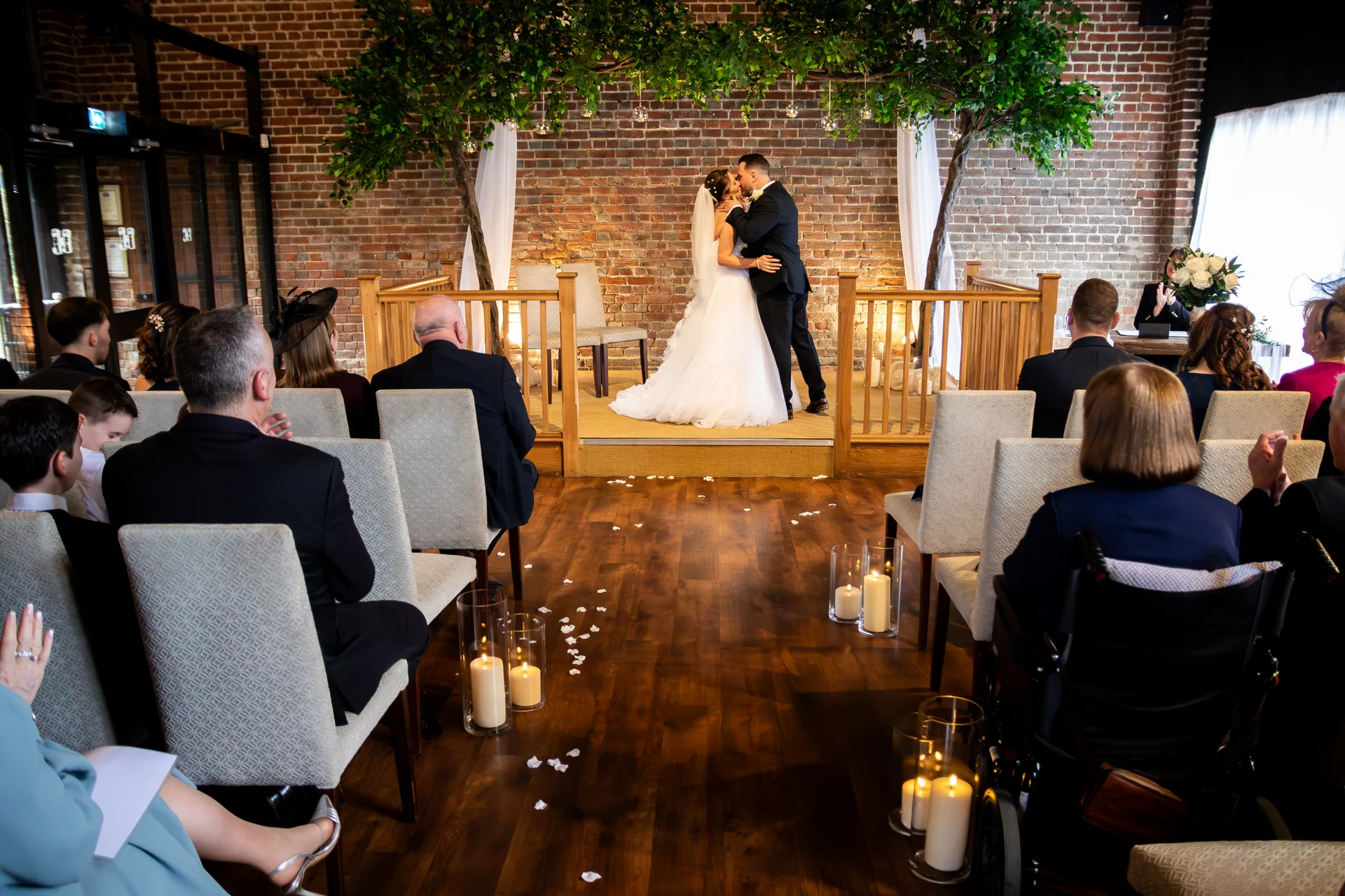 A bride and groom share their first kiss at the altar of an indoor brick venue decorated with tree arches and floor candles while guests watch from seated rows.