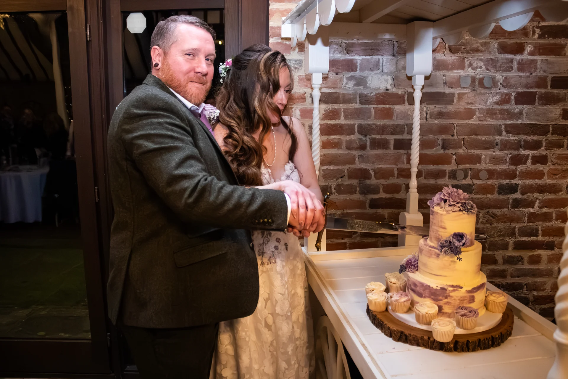 A bride and groom cut a three-tier wedding cake decorated with purple floral accents, standing together in front of an exposed brick wall.