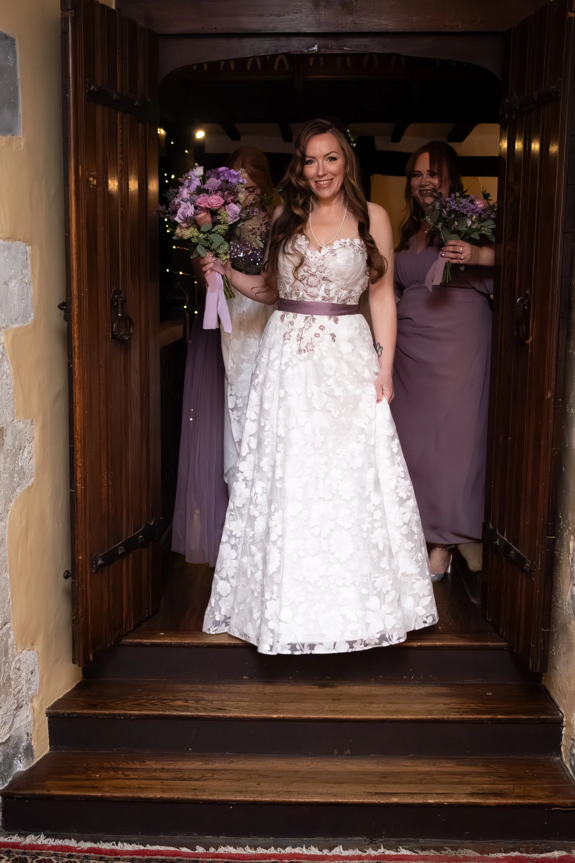 A smiling bride wearing an A-line gown with floral appliqués and a purple sash steps through historic wooden double doors, flanked by bridesmaids holding bouquets.