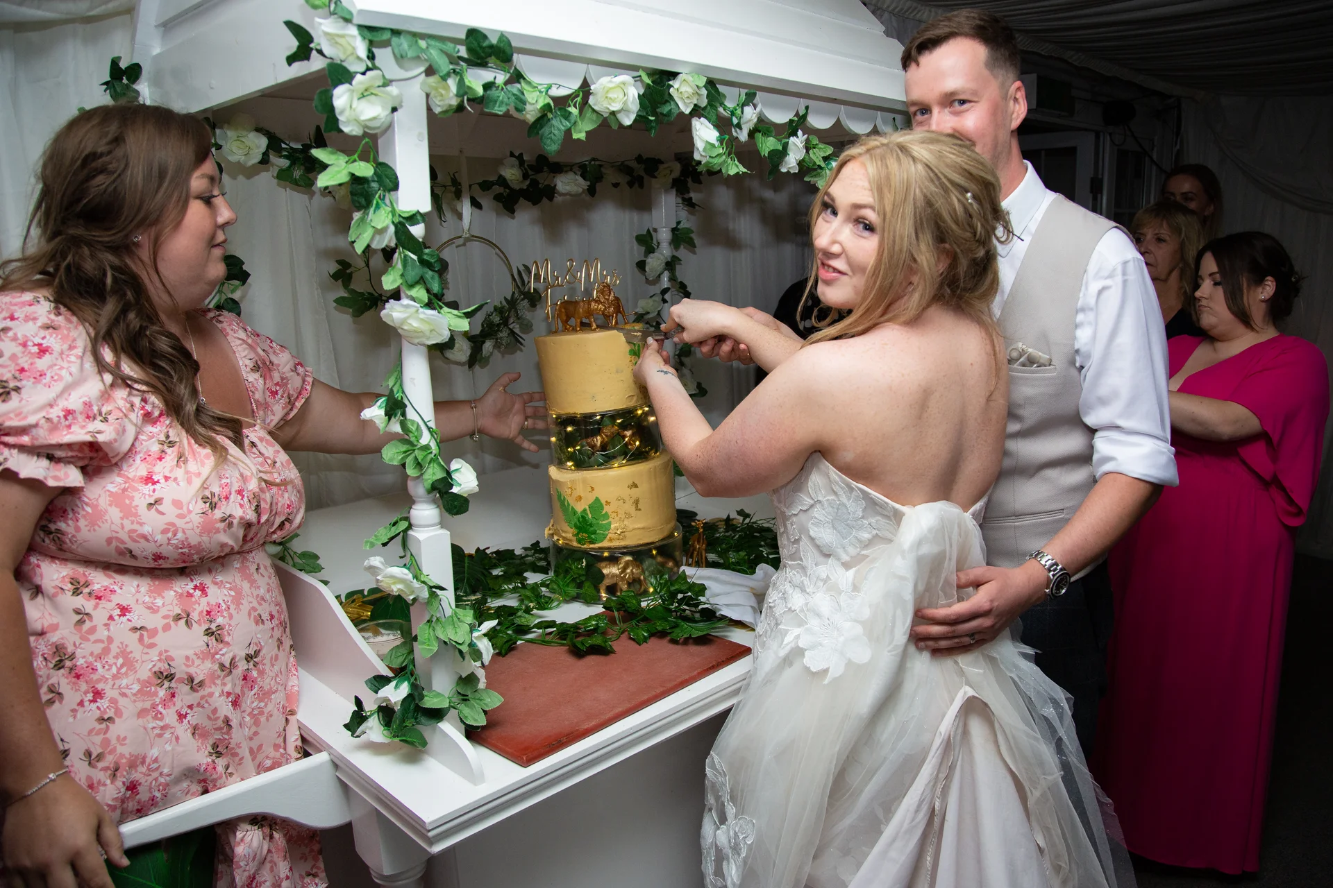 The bride and groom smile over their shoulders while cutting a multi-tiered gold wedding cake displayed on a white floral cart.