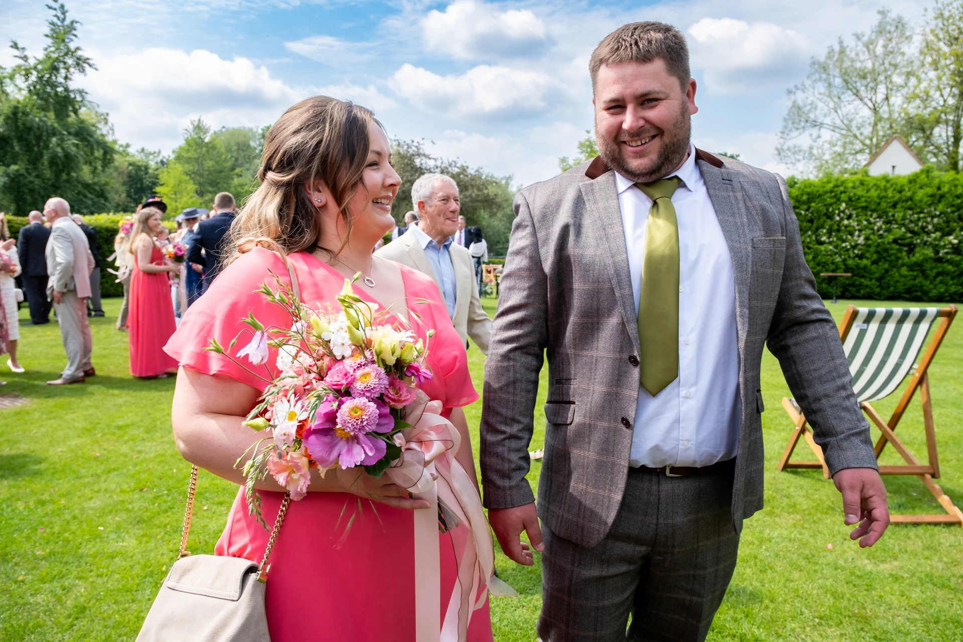A smiling couple walks hand-in-hand across a sunny lawn during an outdoor wedding reception, with the bride holding a colourful wild bouquet and wearing a pink dress, and the groom in a checked suit with a green tie.