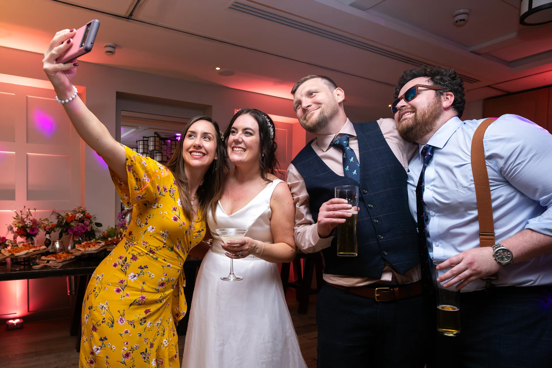 A bride in a white dress and wedding guests take a selfie together at an indoor reception while holding drinks.