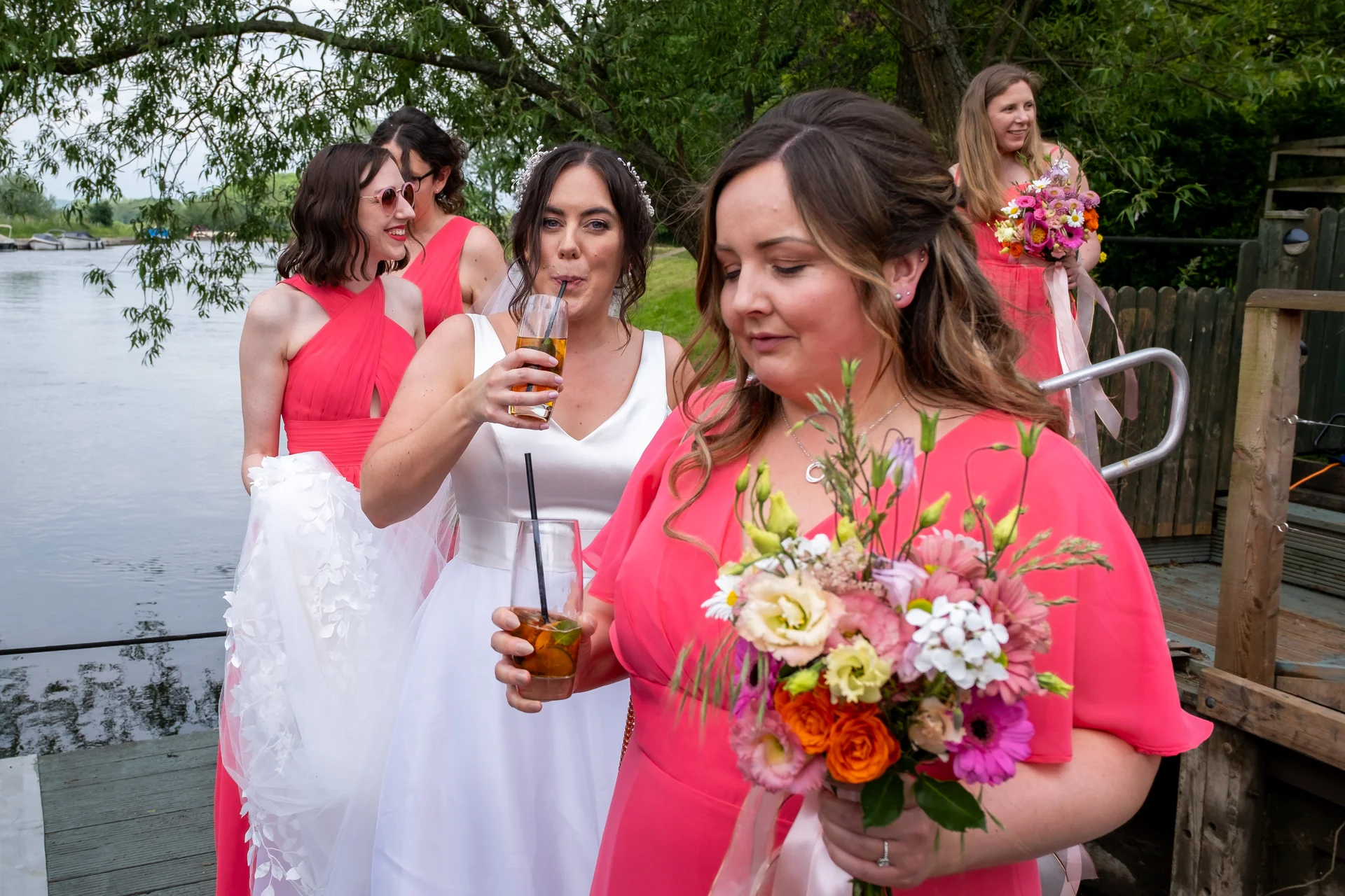 A smiling bride holding a colourful hand-tied bouquet stands alongside her bridesmaids wearing bright pink dresses by a river during a drinks reception.