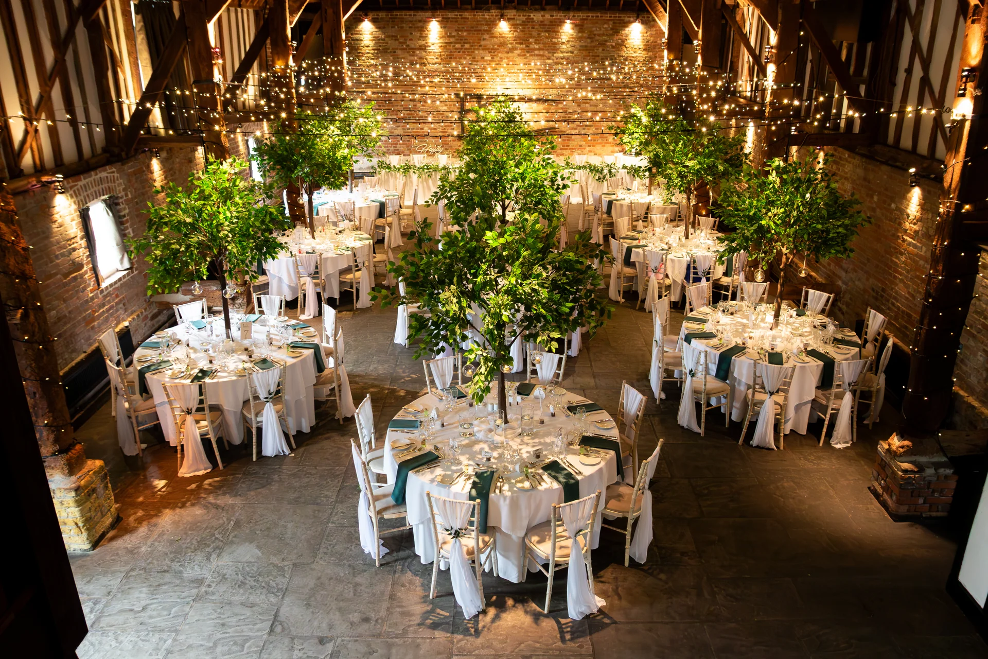 An elevated view of a rustic brick barn wedding reception set up with round tables adorned with white linens, forest green accents, and indoor trees beneath a dazzling canopy of twinkling fairy lights.