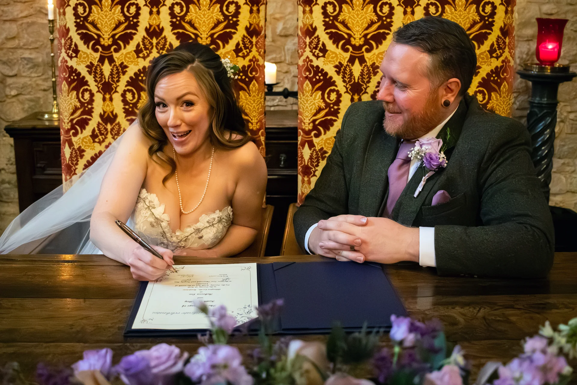 A newlywed bride enthusiastically signs the register while looking back at her groom, who smiles lovingly at her during their wedding ceremony.