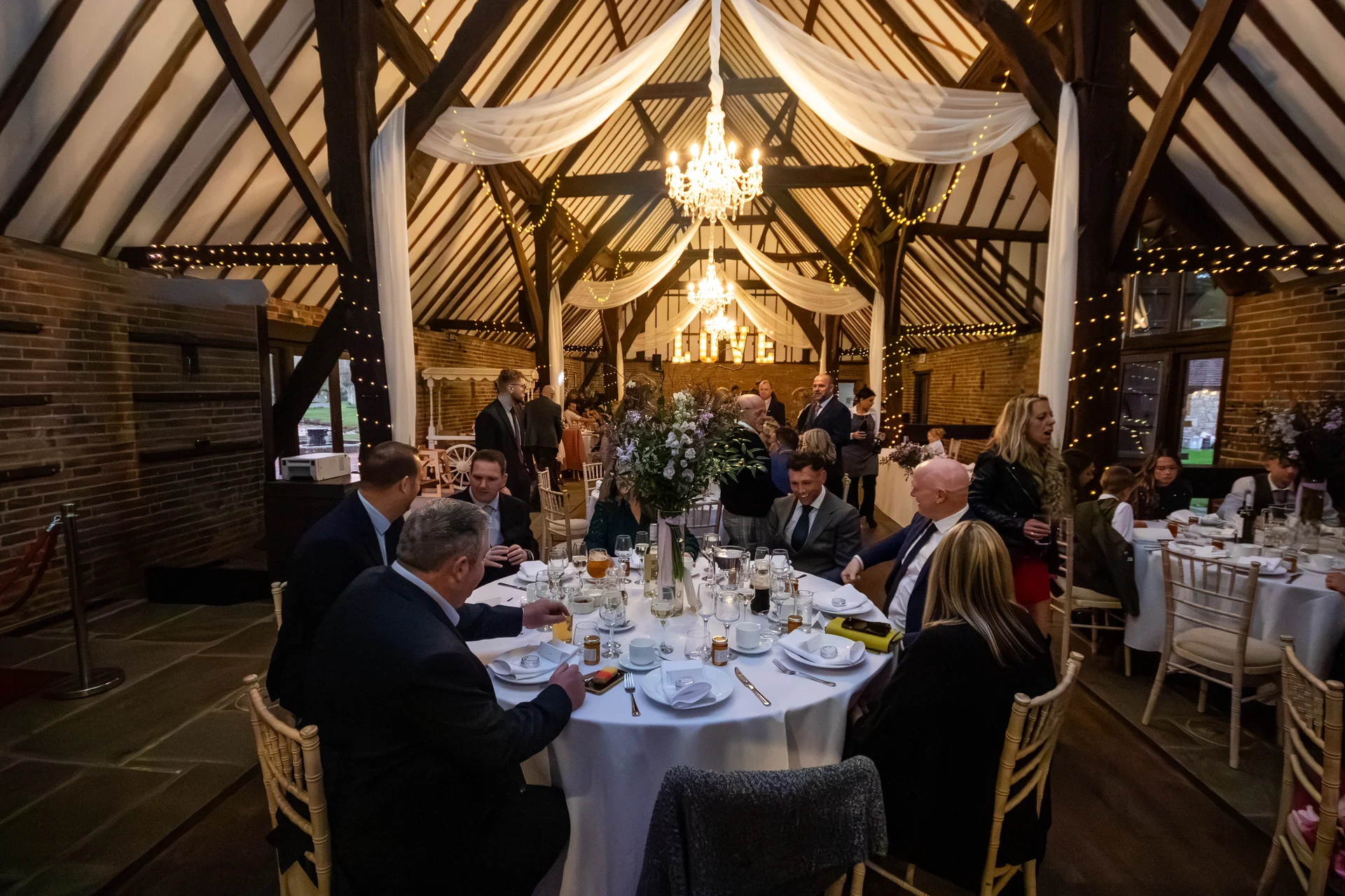 Guests enjoy a wedding breakfast inside a rustic oak-framed barn featuring exposed rafters, twinkling fairy lights, and elegant round dining tables.