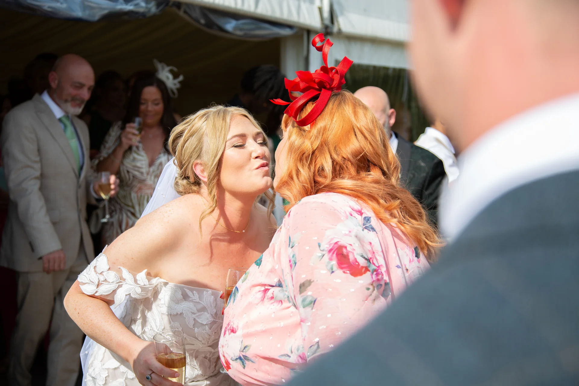 A bride in an off-the-shoulder gown leans in to greet a guest wearing a pink floral dress and a striking red fascinator during a sunny wedding reception.