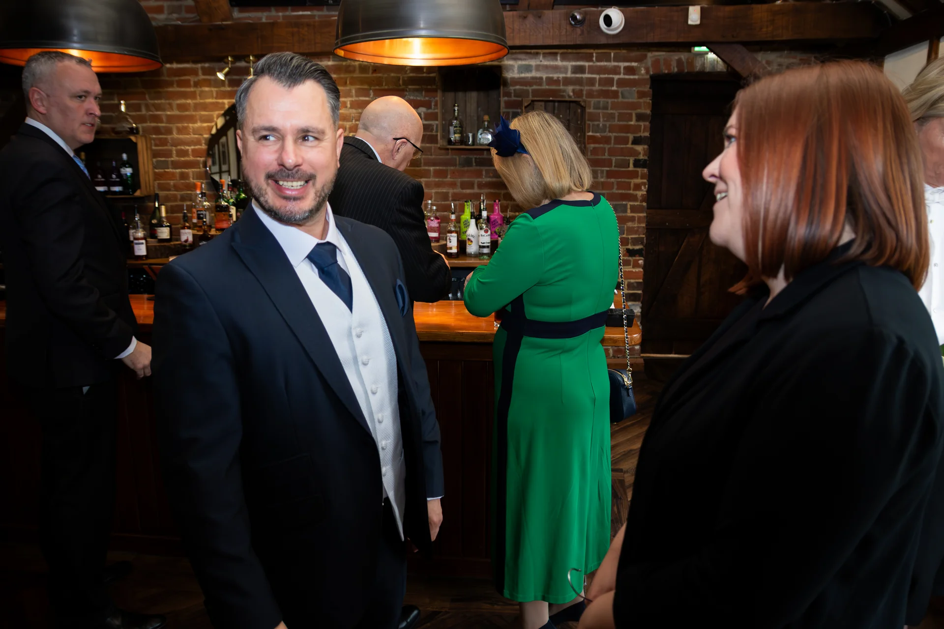 Wedding guests mingle and chat near a wooden bar inside a venue with exposed brick walls and warm pendant lighting.