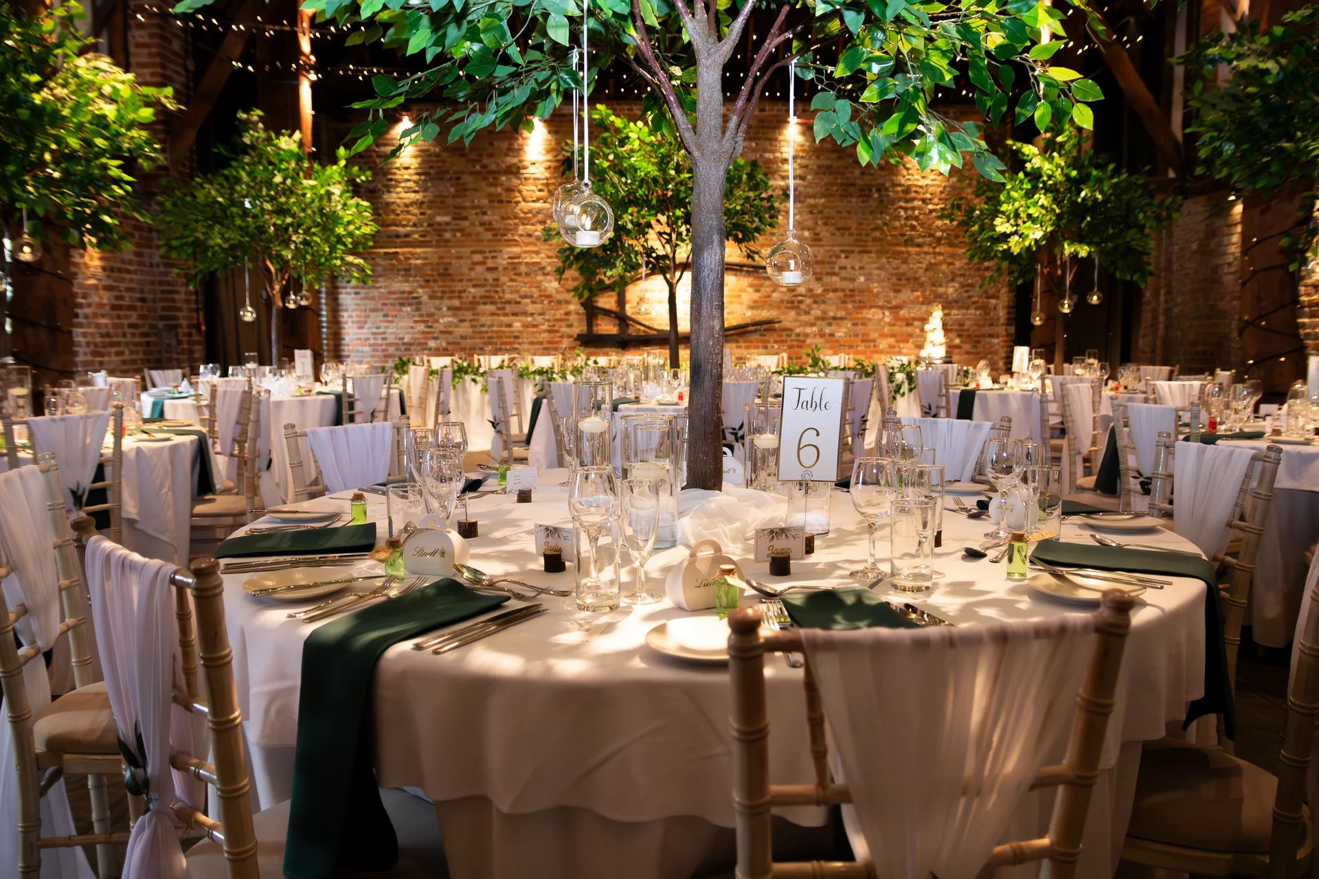 A beautifully styled round banquet table in a rustic barn featuring exposed brick walls, emerald green napkins, and a central decorative tree with hanging glass baubles.