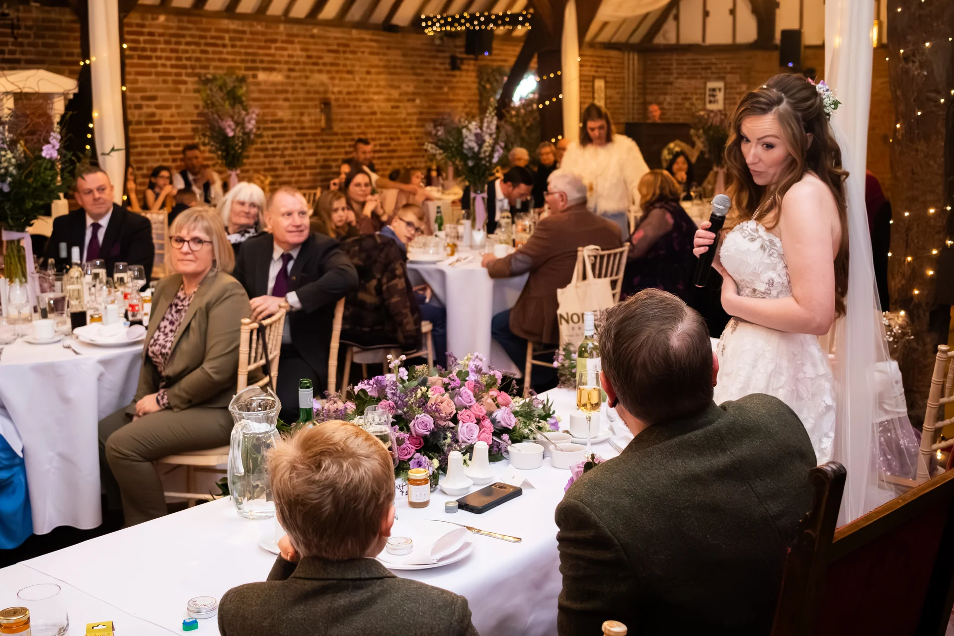 A bride gives a speech with a microphone at the top table during a rustic barn wedding reception, with seated guests looking on attentively.