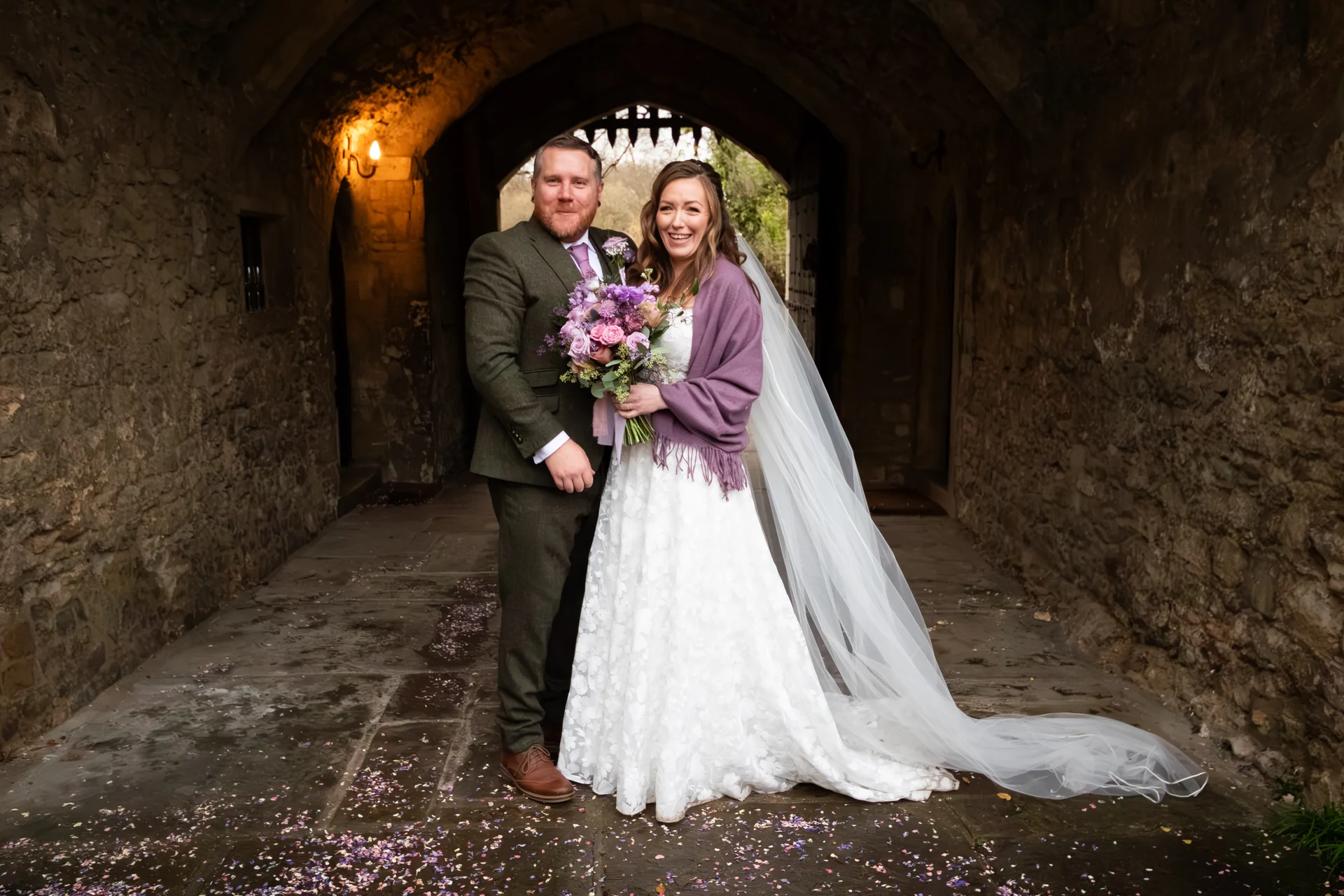 A newlywed couple poses together inside a stone archway, with the bride holding a vibrant bouquet and wearing a flowing white lace gown with a cathedral veil.
