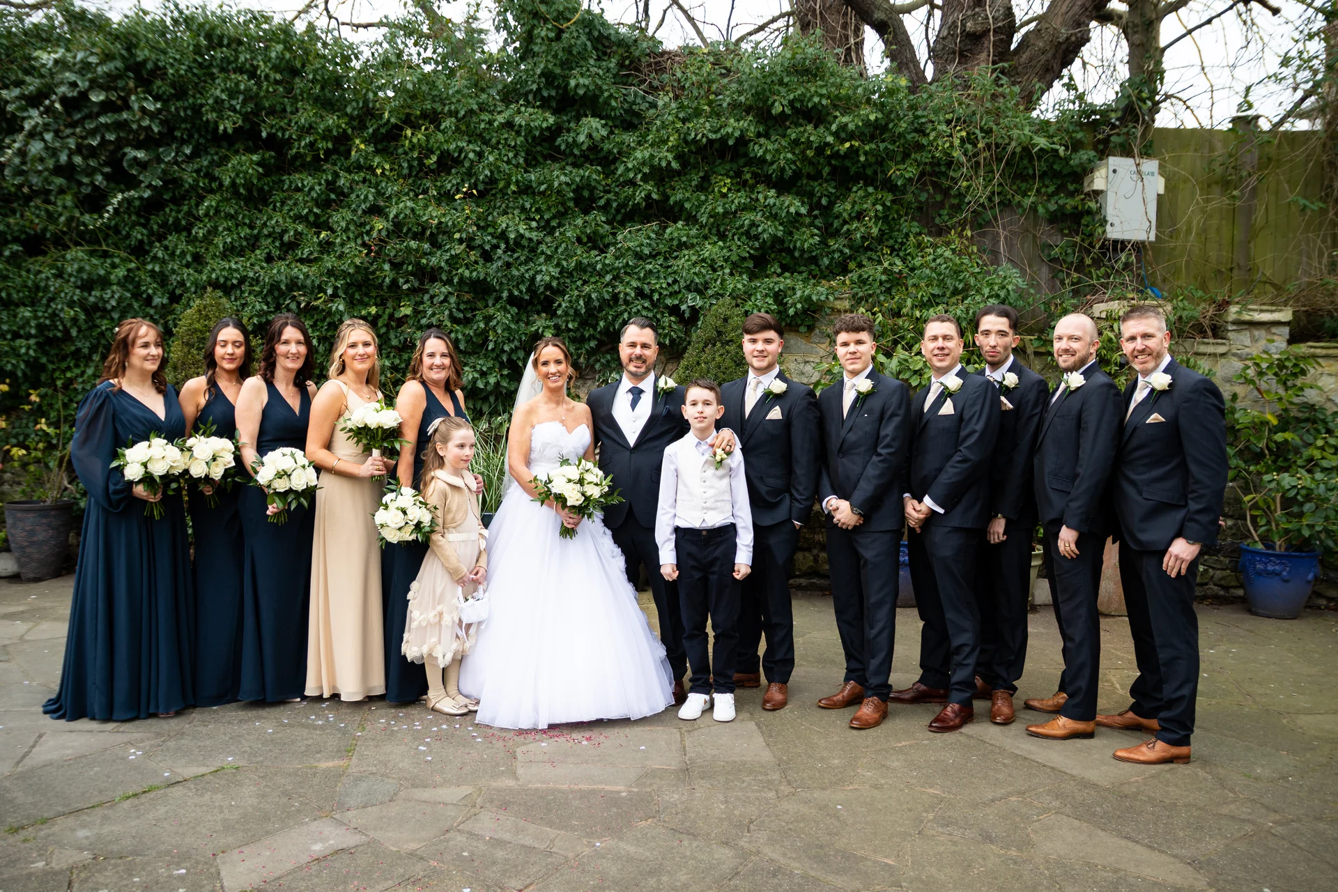 A full-length group portrait of the bride, groom, bridesmaids, and groomsmen posing together outdoors in front of a lush ivy-covered wall on a paved courtyard.