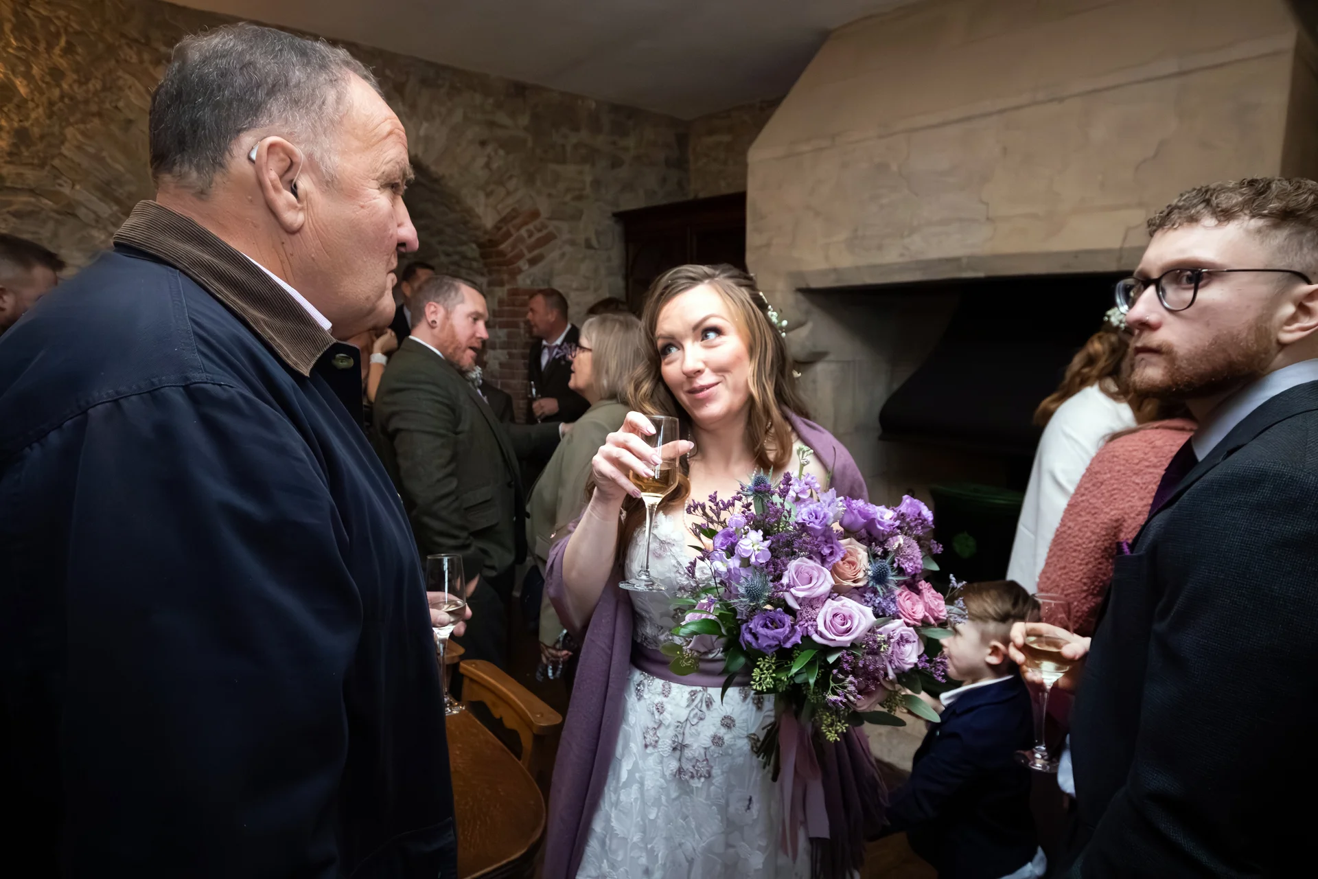 A smiling bride holding a purple floral bouquet and a glass of champagne, surrounded by guests mingling inside a rustic stone venue with a large fireplace.