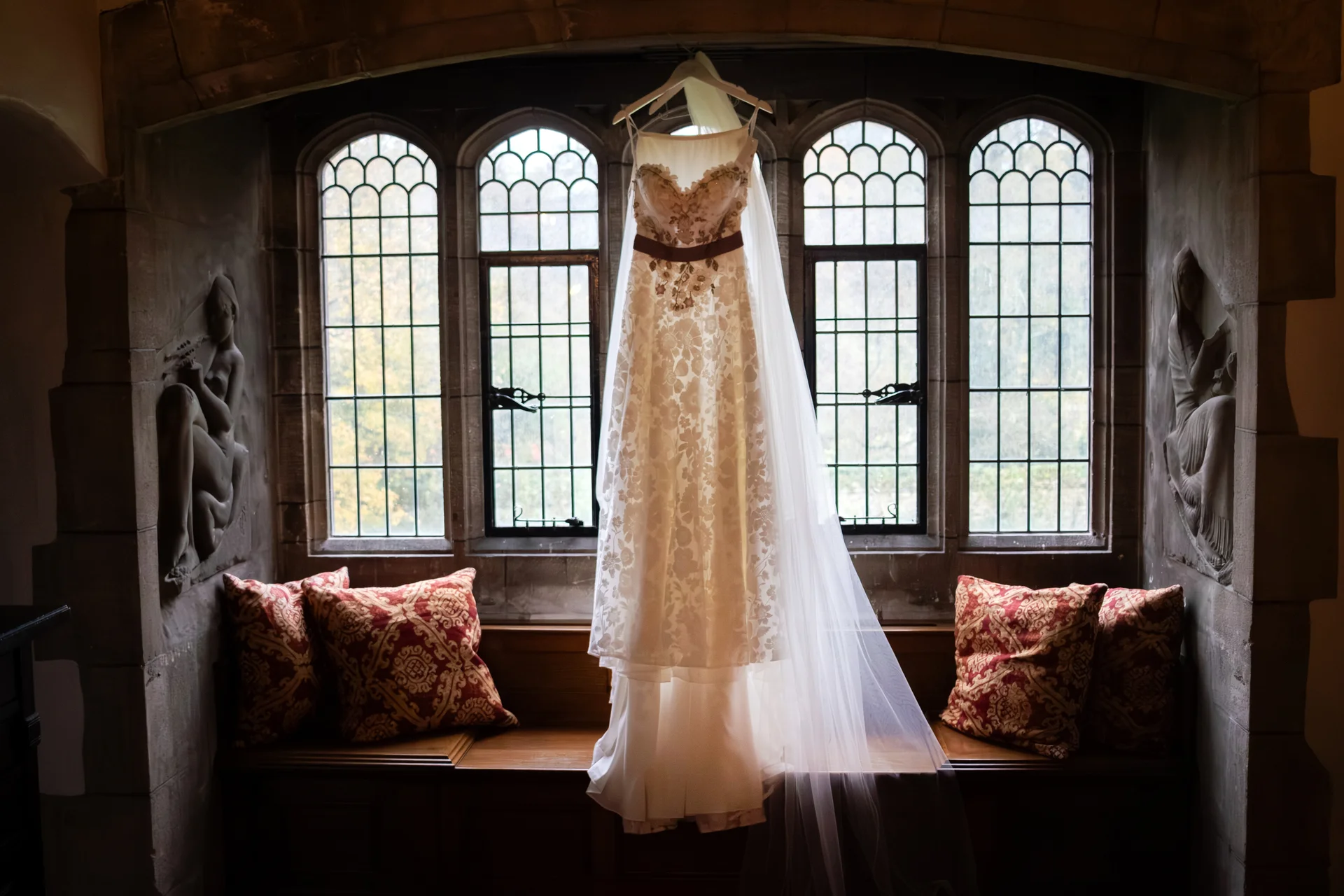 An intricately patterned lace wedding dress hangs in front of a grand arched stone window with stained glass panes, accompanied by a flowing veil and decorative cushions.