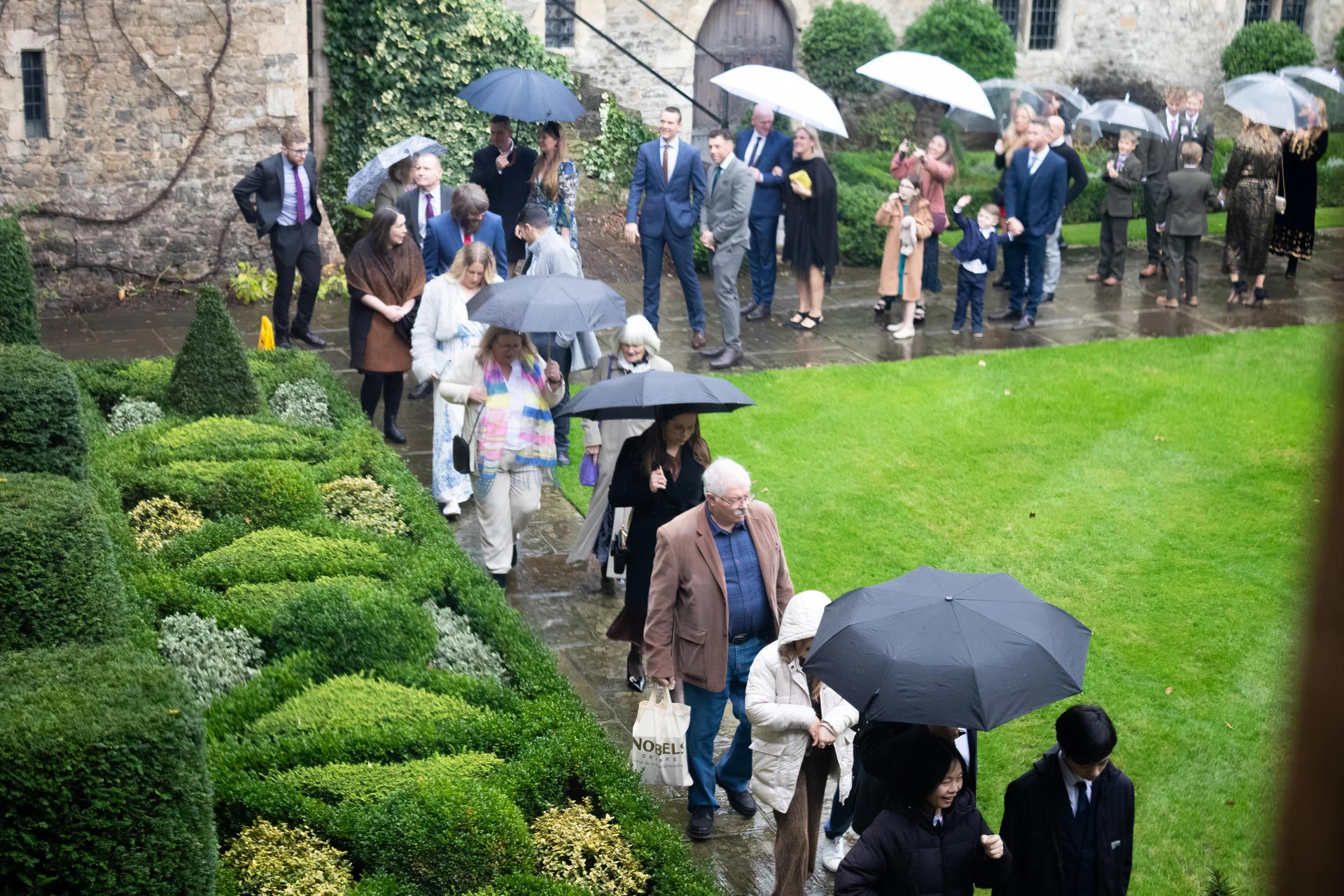 Guests walk along a damp stone pathway past manicured garden hedges holding umbrellas during a rainy wedding day.