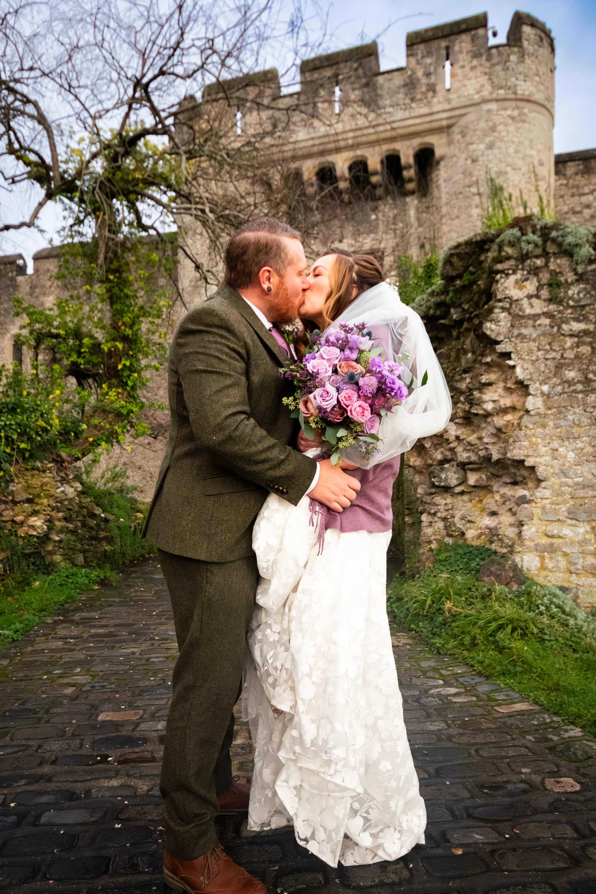 A newlywed couple shares a tender kiss on a cobbled path outside an ancient stone castle, with the bride holding a vibrant purple bouquet.