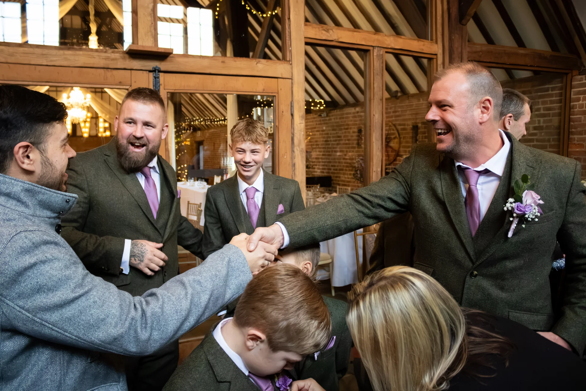 A group of cheerful guests and groomsmen in green tweed suits and purple ties share a joyful moment of laughter and handshaking inside a rustic barn venue.