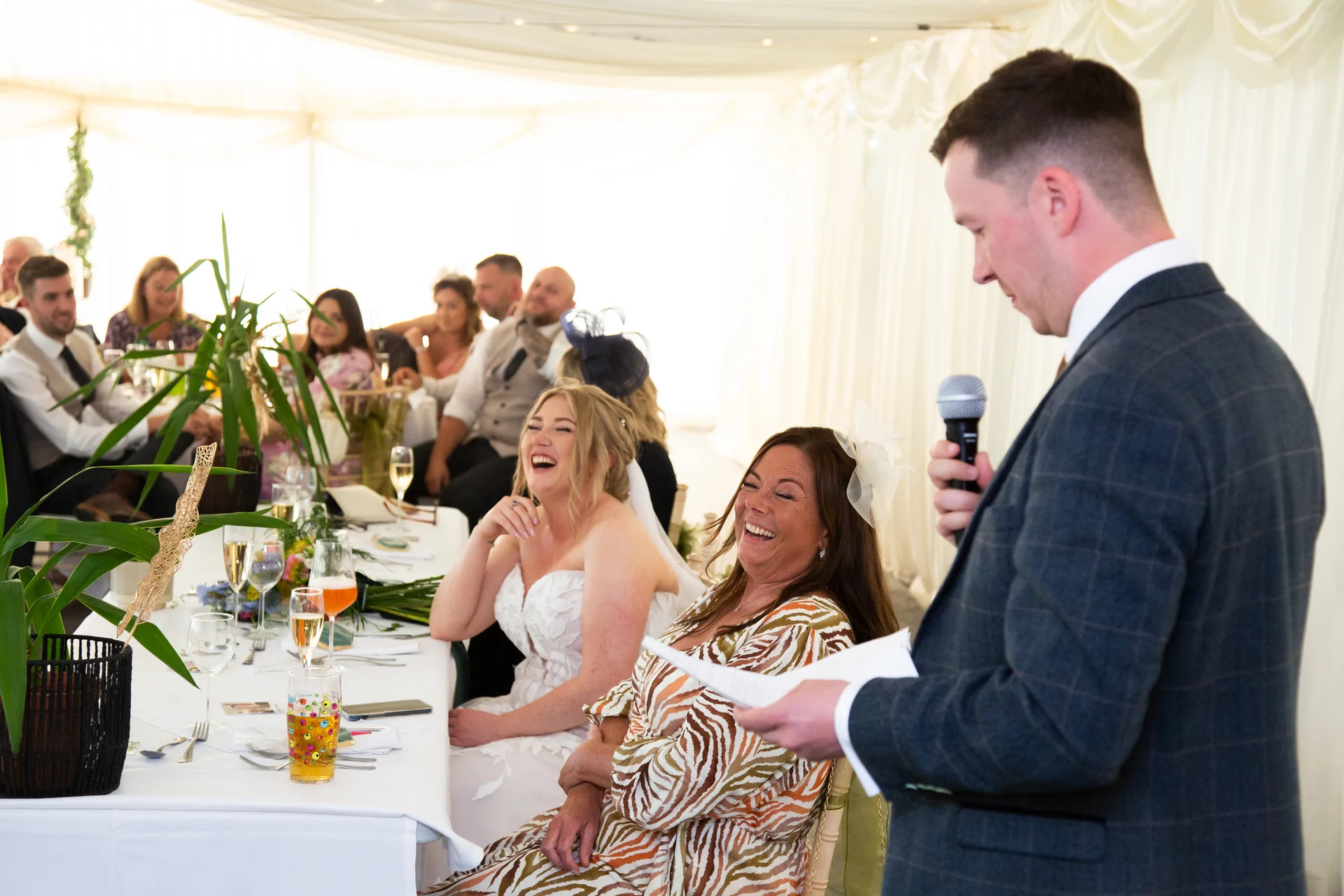 A groom delivers a speech in a marquee, causing the bride and a seated female guest to burst into laughter.