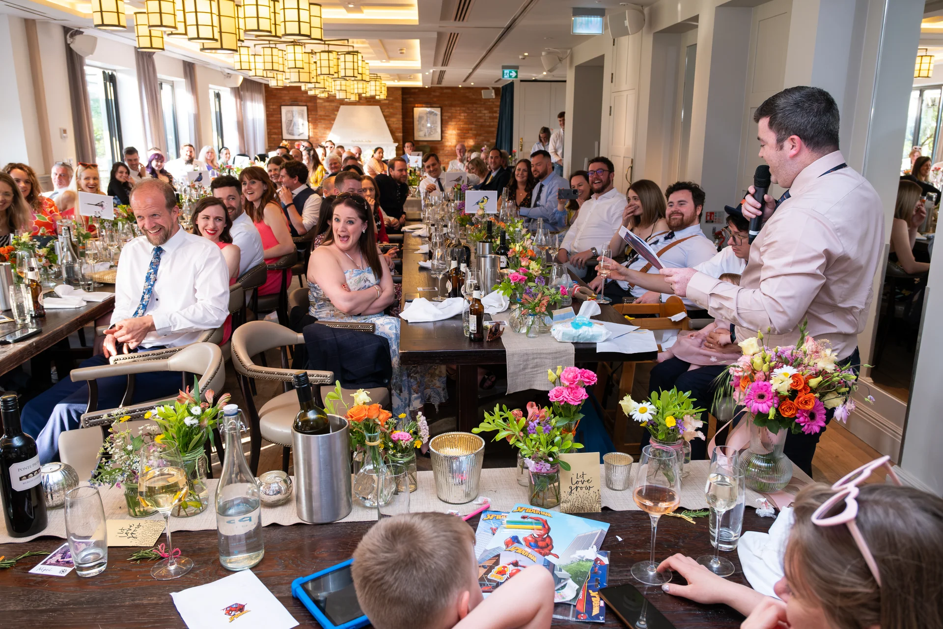 A wedding guest stands and delivers a speech to a room full of seated, laughing guests gathered around long banquet tables.
