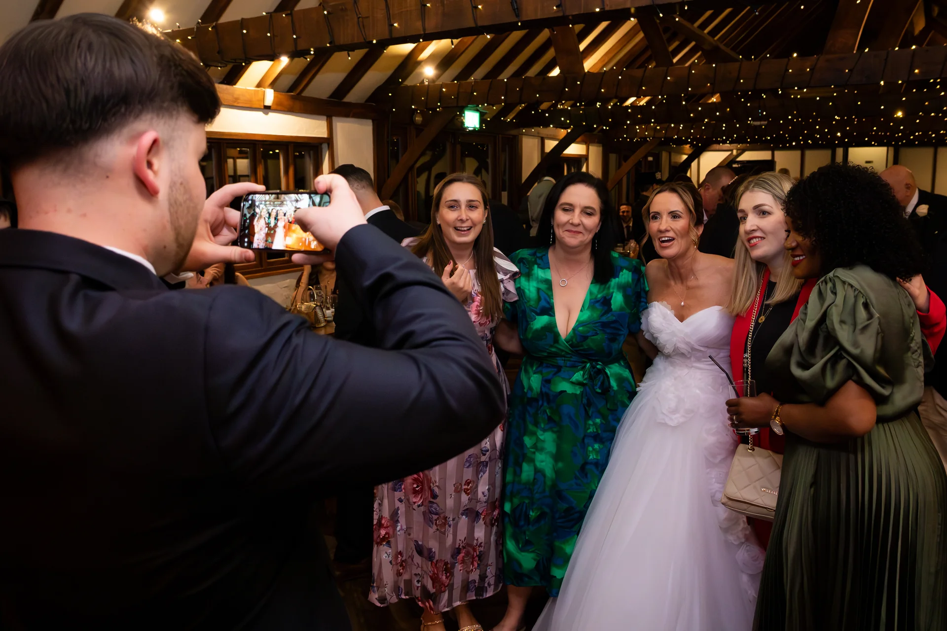 A wedding guest takes a photo with his smartphone of a bride in a strapless tulle gown posing with laughing friends inside a rustic barn illuminated by a fairy light canopy.