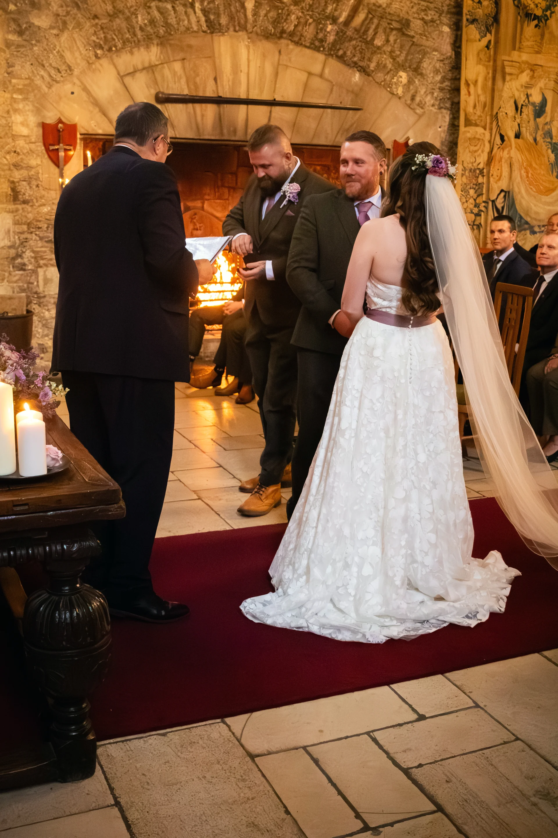A bride in a floral applique gown with a long veil stands with the groom and groomsmen during a candlelit ceremony inside a historic castle venue.