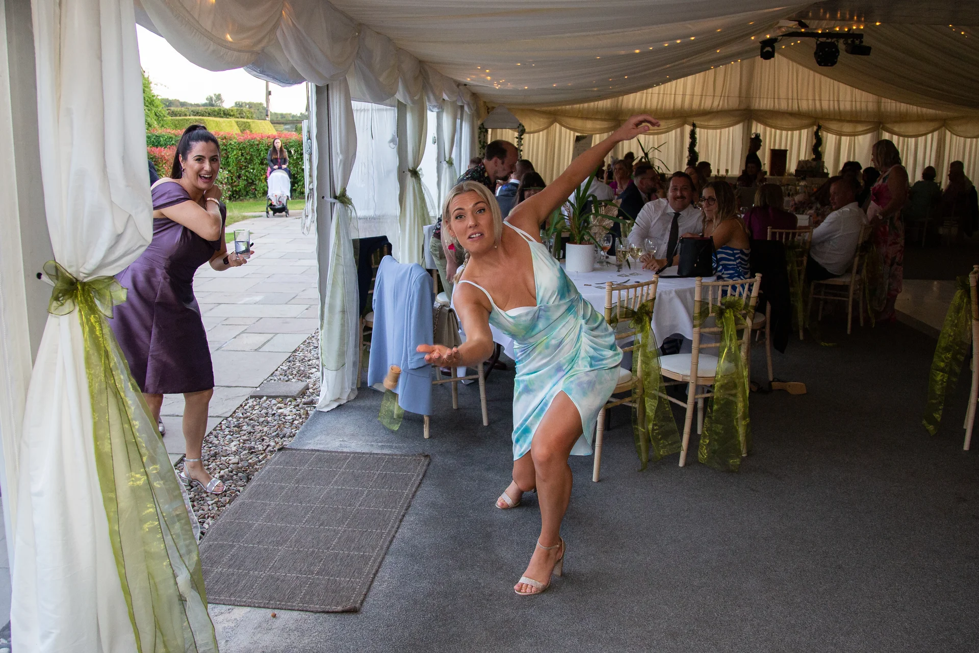 A guest dynamically lurches forward to toss her bouquet inside a white draped marquee during a wedding reception, while onlookers watch and laugh.