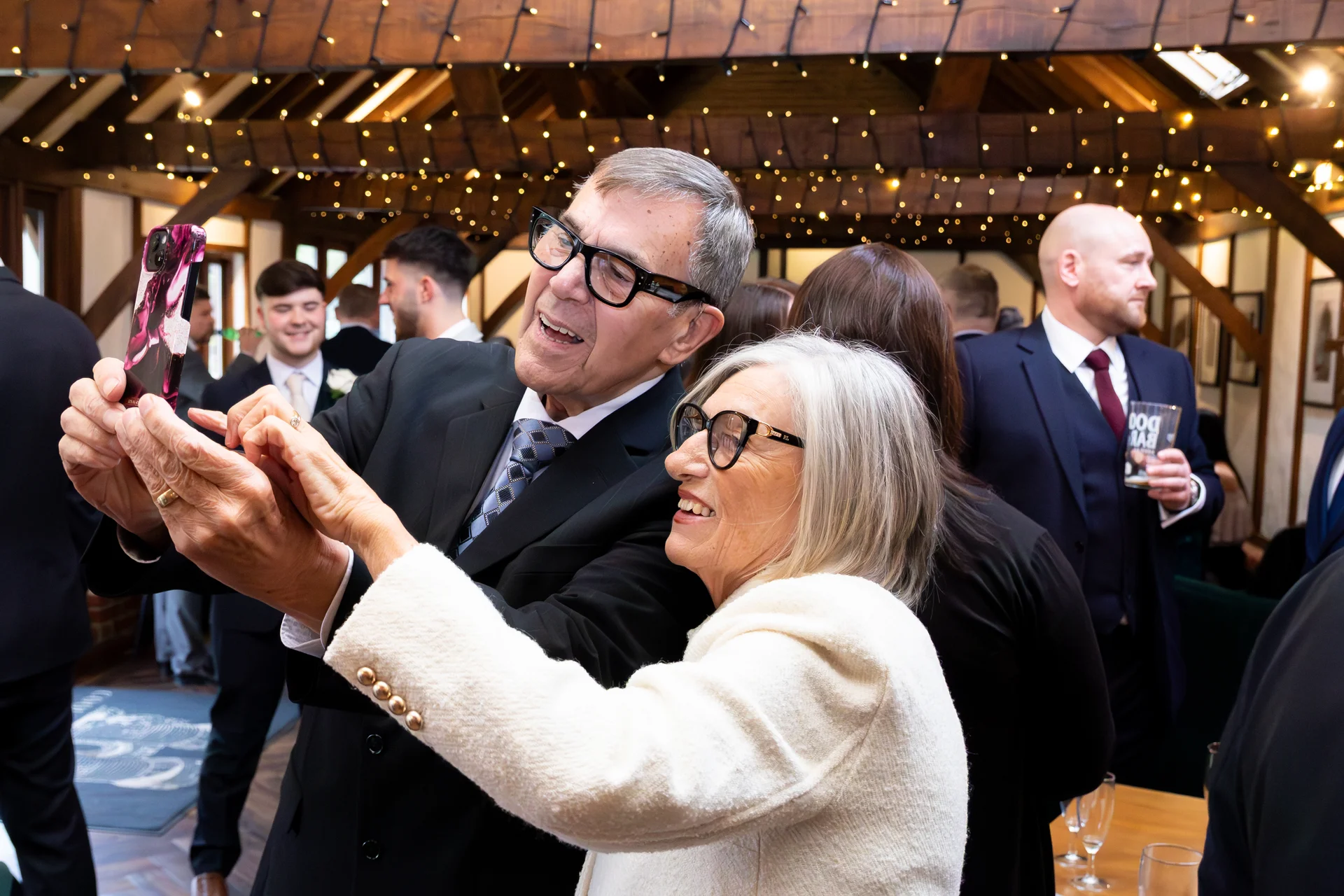 An older couple joyfully takes a selfie together at a wedding reception inside a rustic barn venue with a lit fairy light canopy.
