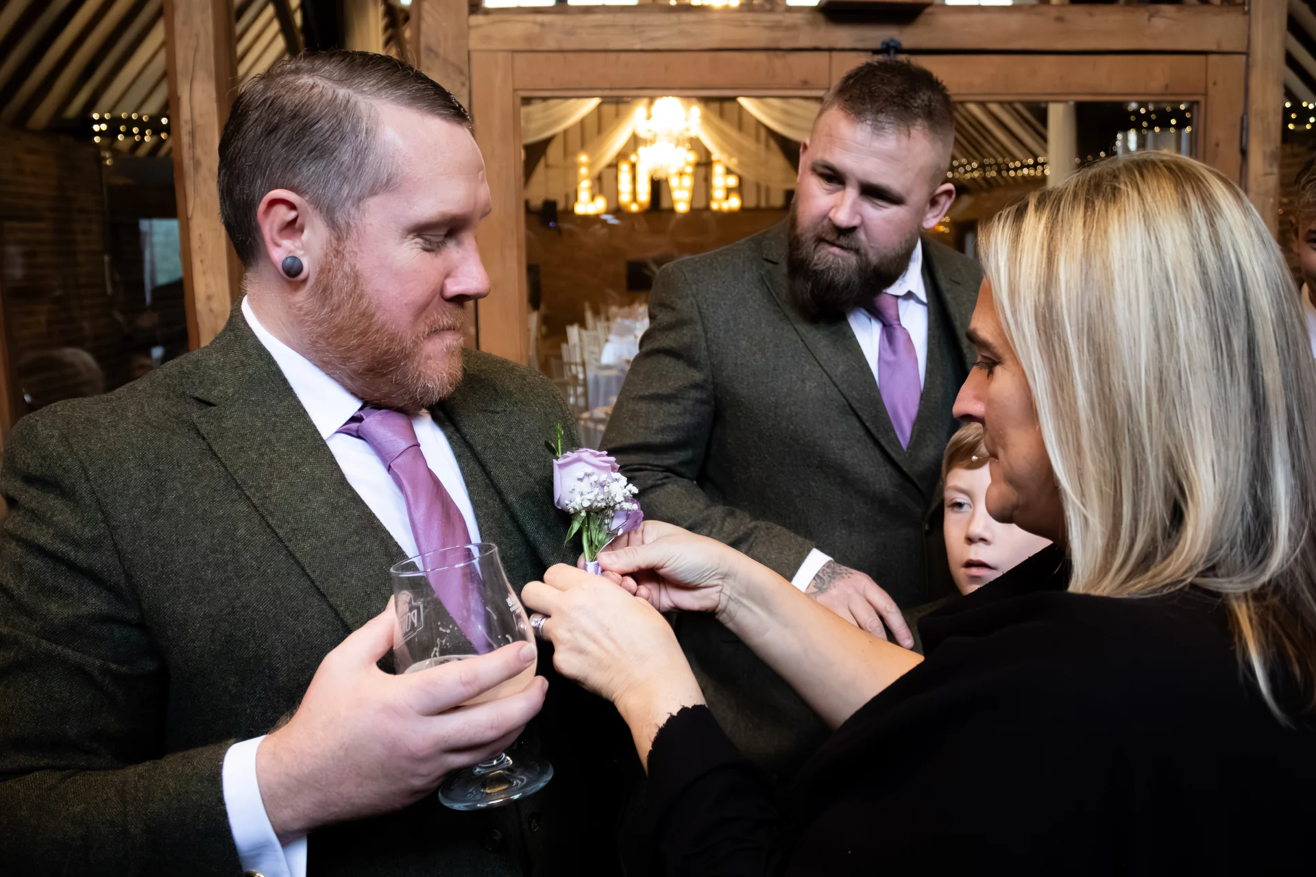 A groom wearing a tweed suit and purple tie gets his floral buttonhole adjusted during wedding preparations inside a rustic barn.