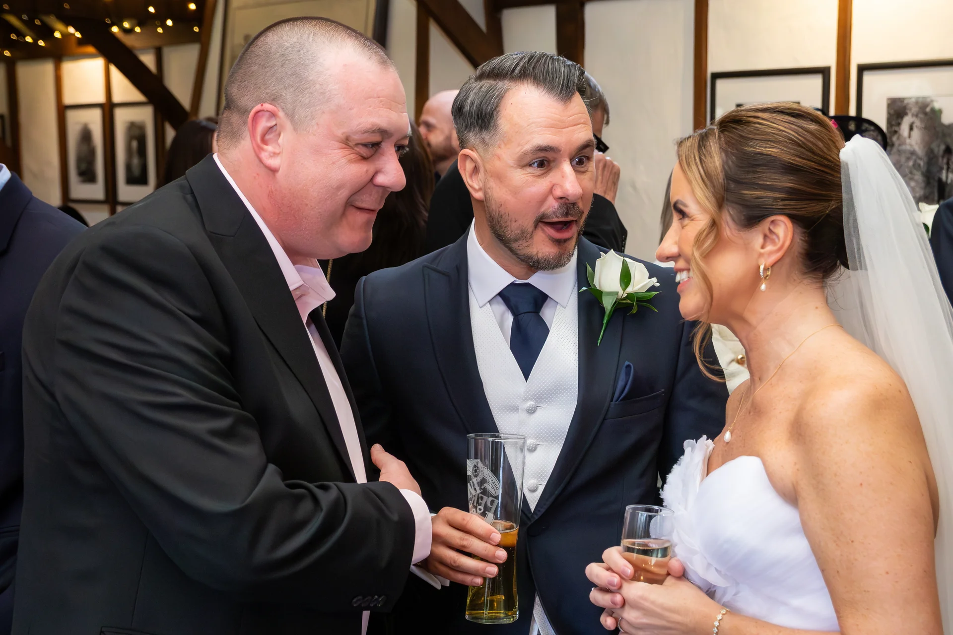 A groom in a dark suit with a white waistcoat and rose buttonhole chats animatedly with a guest holding a pint glass, alongside the smiling bride in a white strapless gown during the wedding reception.