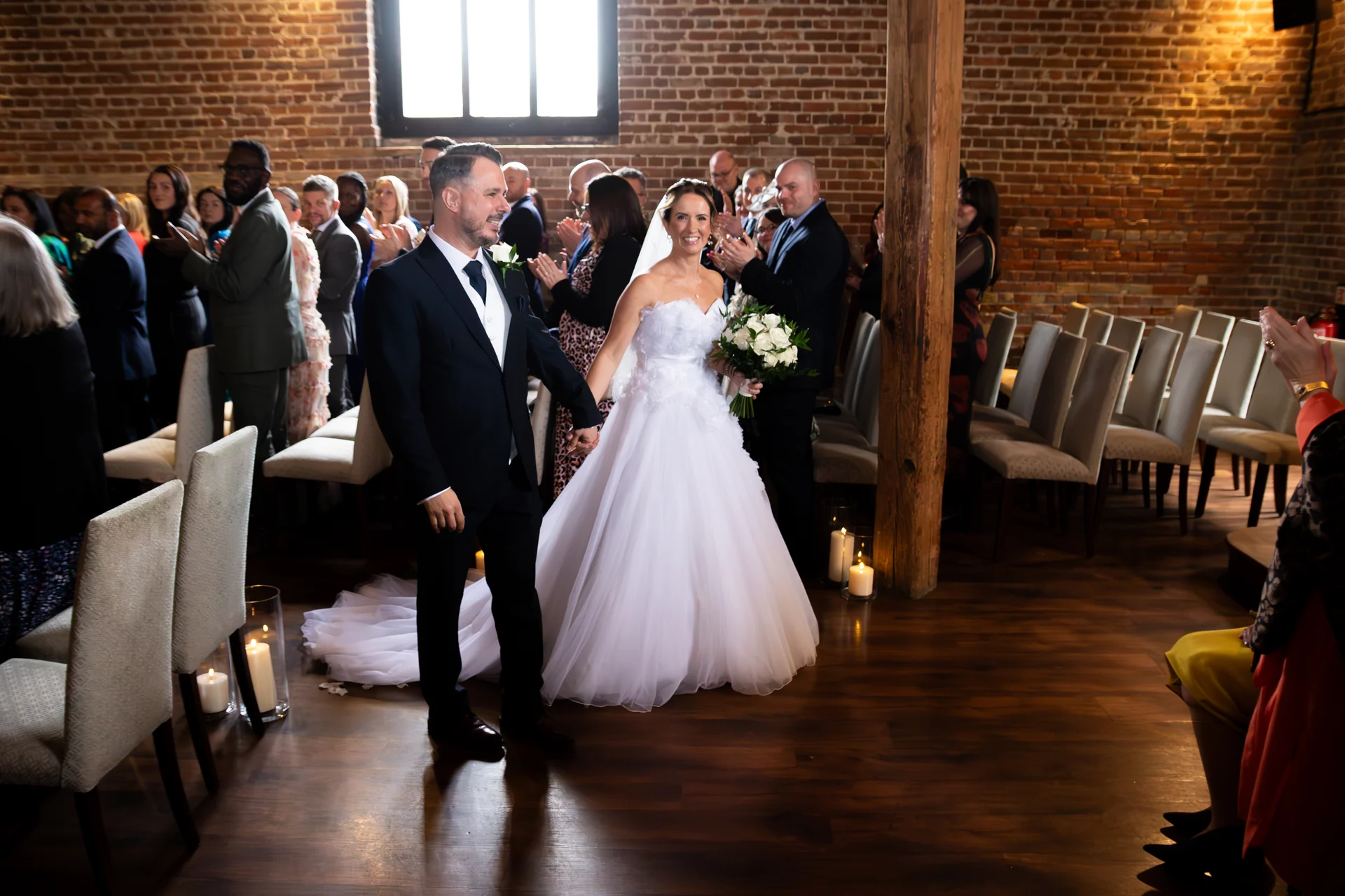 Newlywed bride and groom smiling happily and walking back down the aisle together as guests applaud in a rustic brick barn venue.