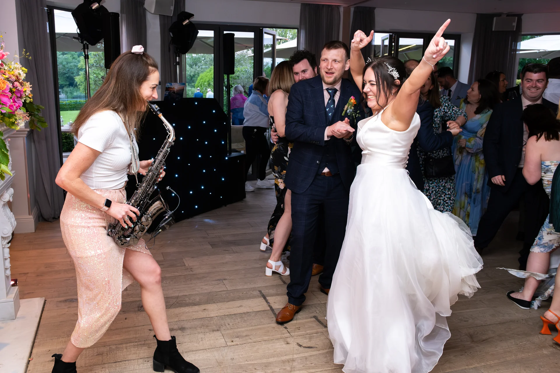 A joyful bride in a white gown dances excitedly on the dance floor alongside her groom while a live saxophonist performs nearby.