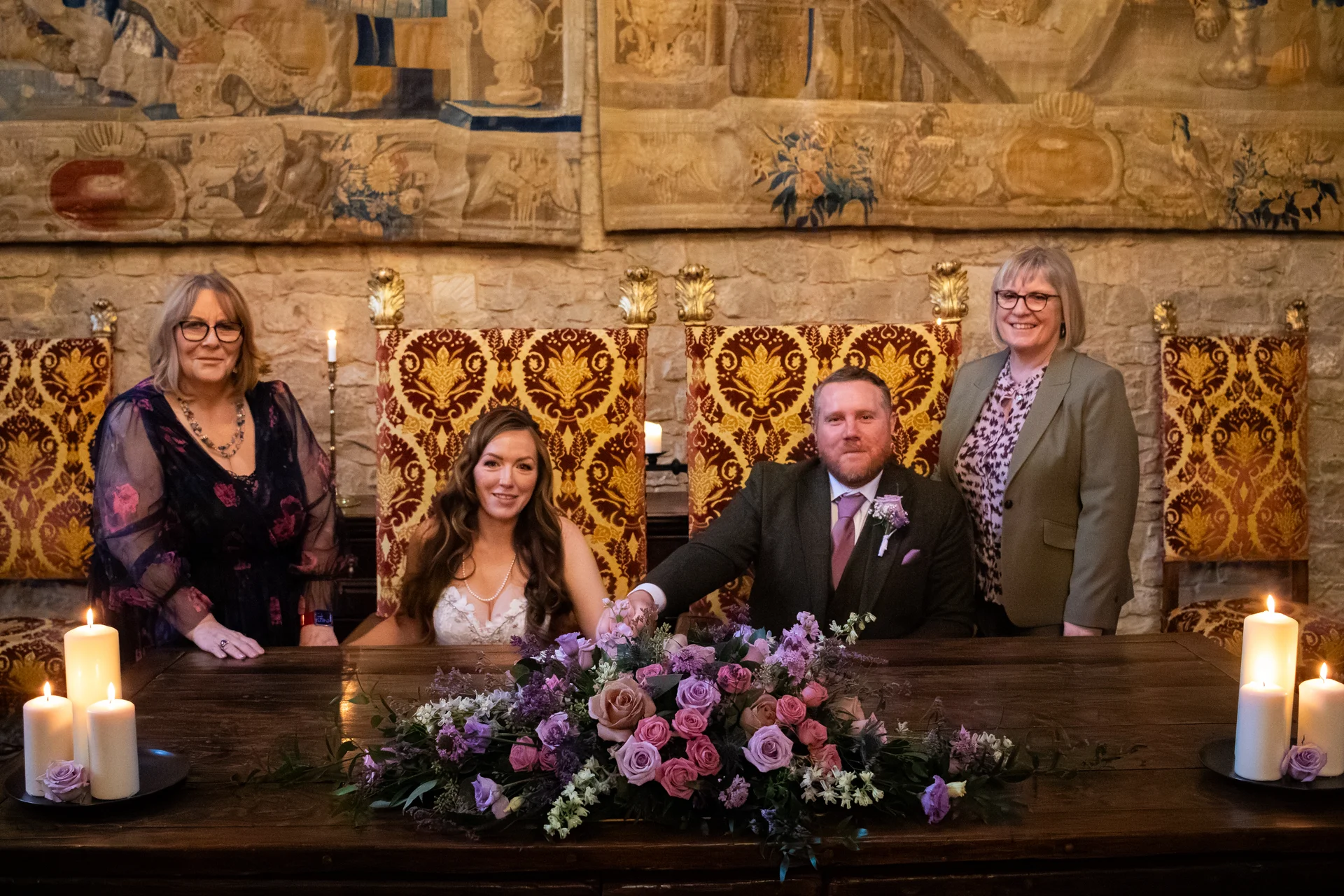 A newly married couple sits together at a rustic wooden table during their civil ceremony, flanked by witnesses and framed by historic stone walls and decorative tapestries.