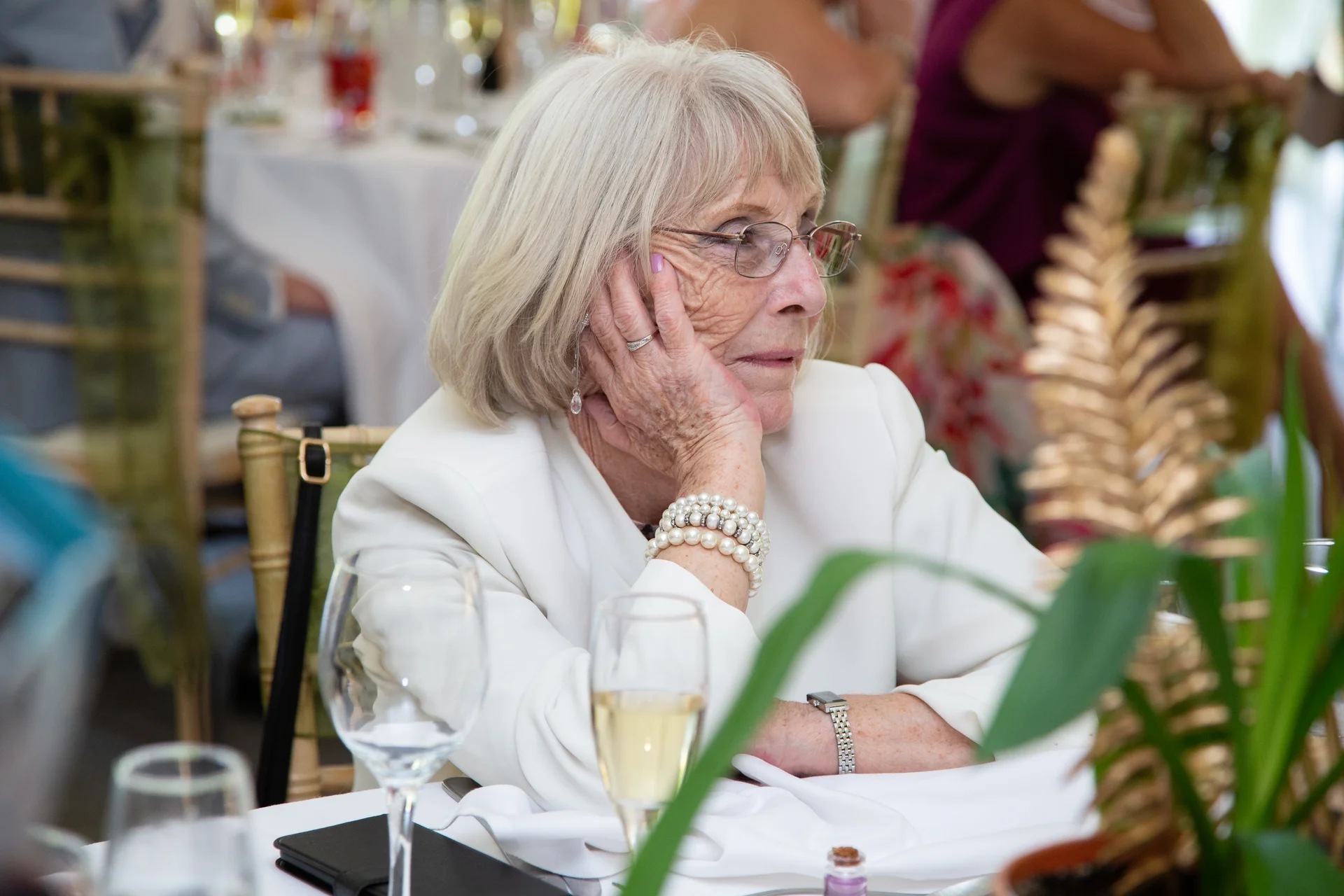 An elderly female wedding guest with silver hair and glasses rests her chin in her hand while seated at a reception table.