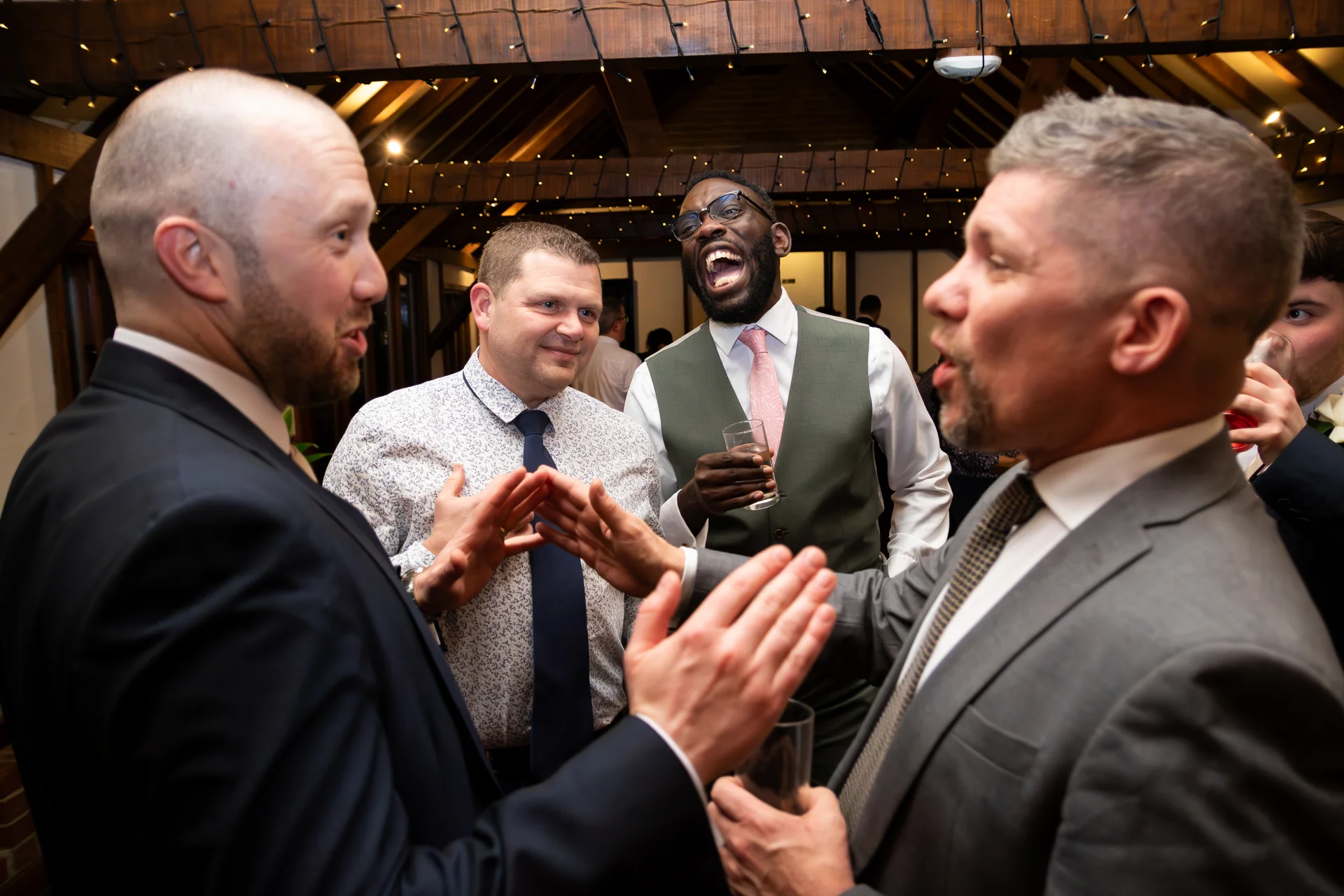 Wedding guests share a joyful, animated conversation and laugh loudly together during an evening reception inside a rustic barn illuminated by fairy lights.