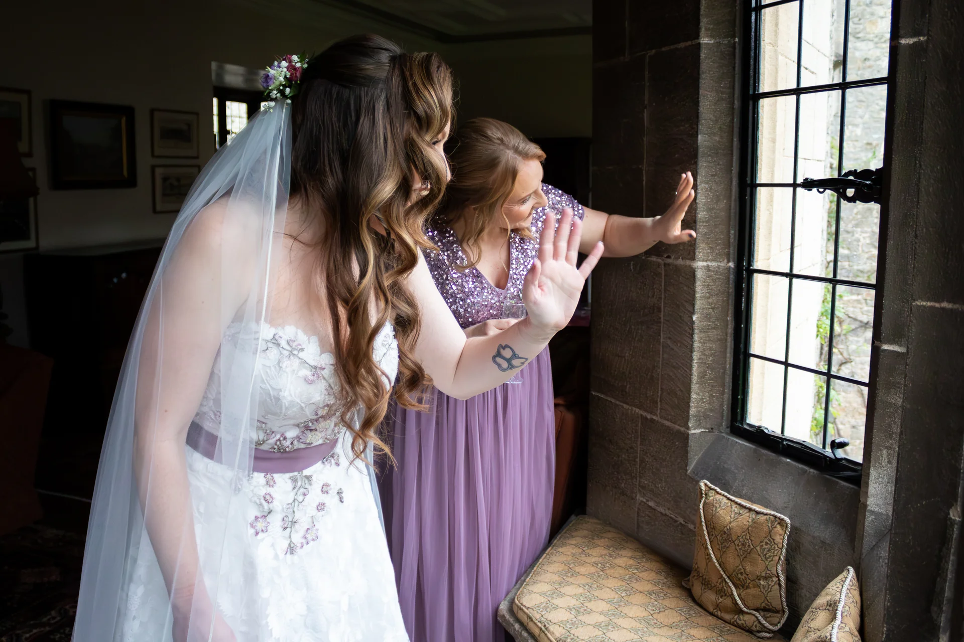 A bride in a white gown with a purple sash and floral appliques looks out of a stone framed window alongside a bridesmaid wearing a sparkly purple dress.