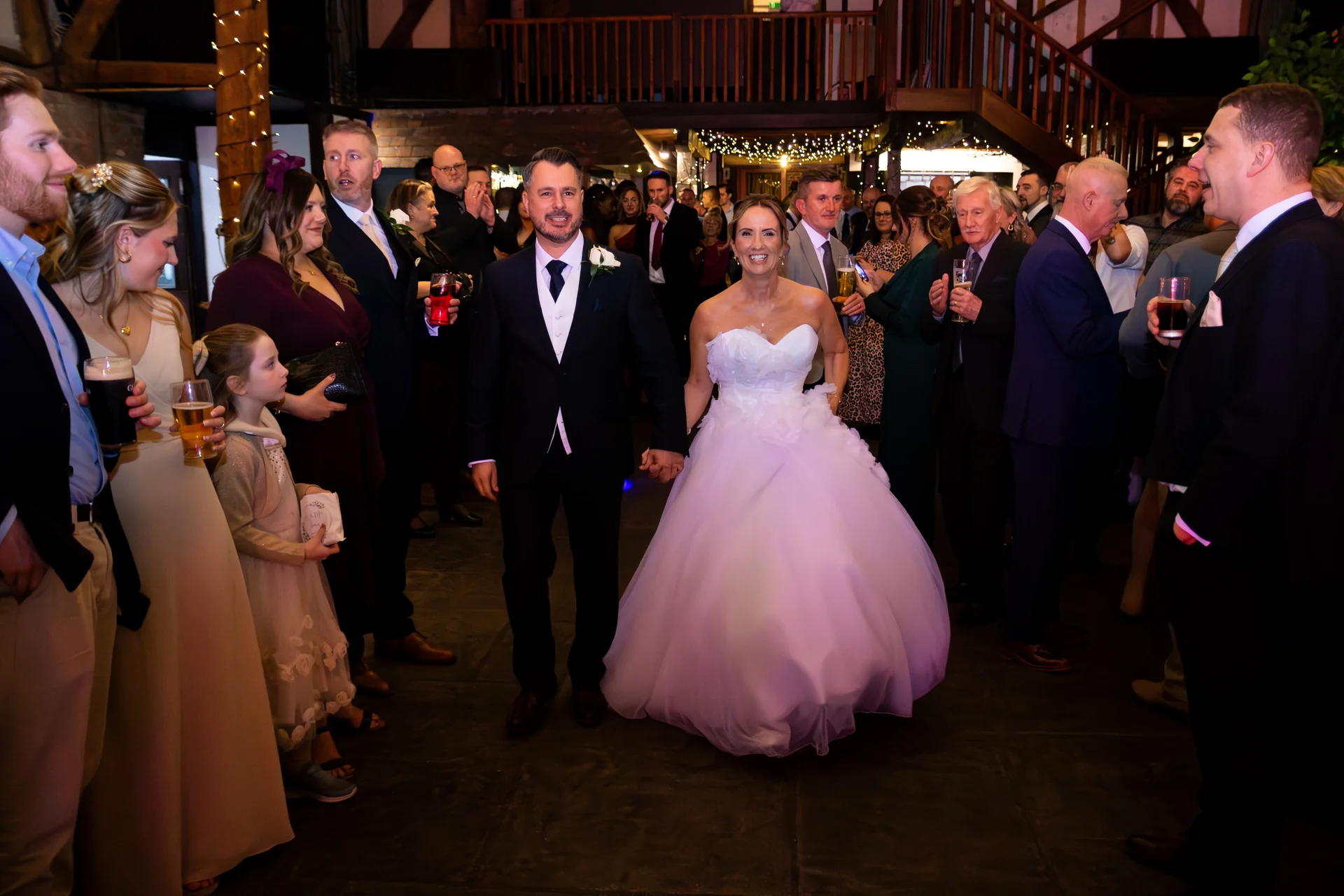 Newlyweds smiling happily as they enter their rustic barn evening reception, holding hands while surrounded by cheering guests holding drinks.