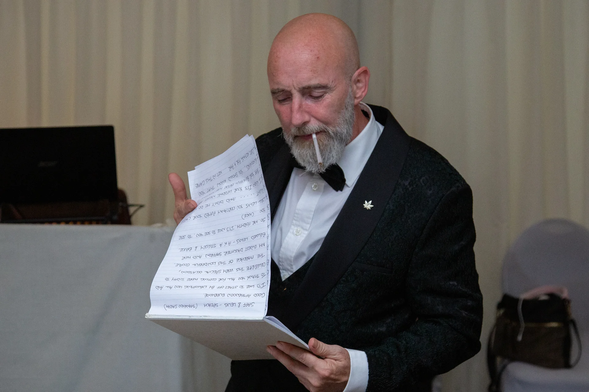A mature man in a black tuxedo and bow tie holds handwritten wedding speech notes while speaking.