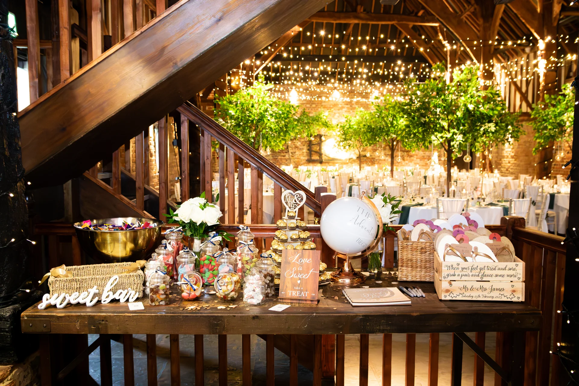 A rustic barn wedding reception features a wooden table styled as a sweet bar and guest book station, illuminated by a warm fairy light canopy.