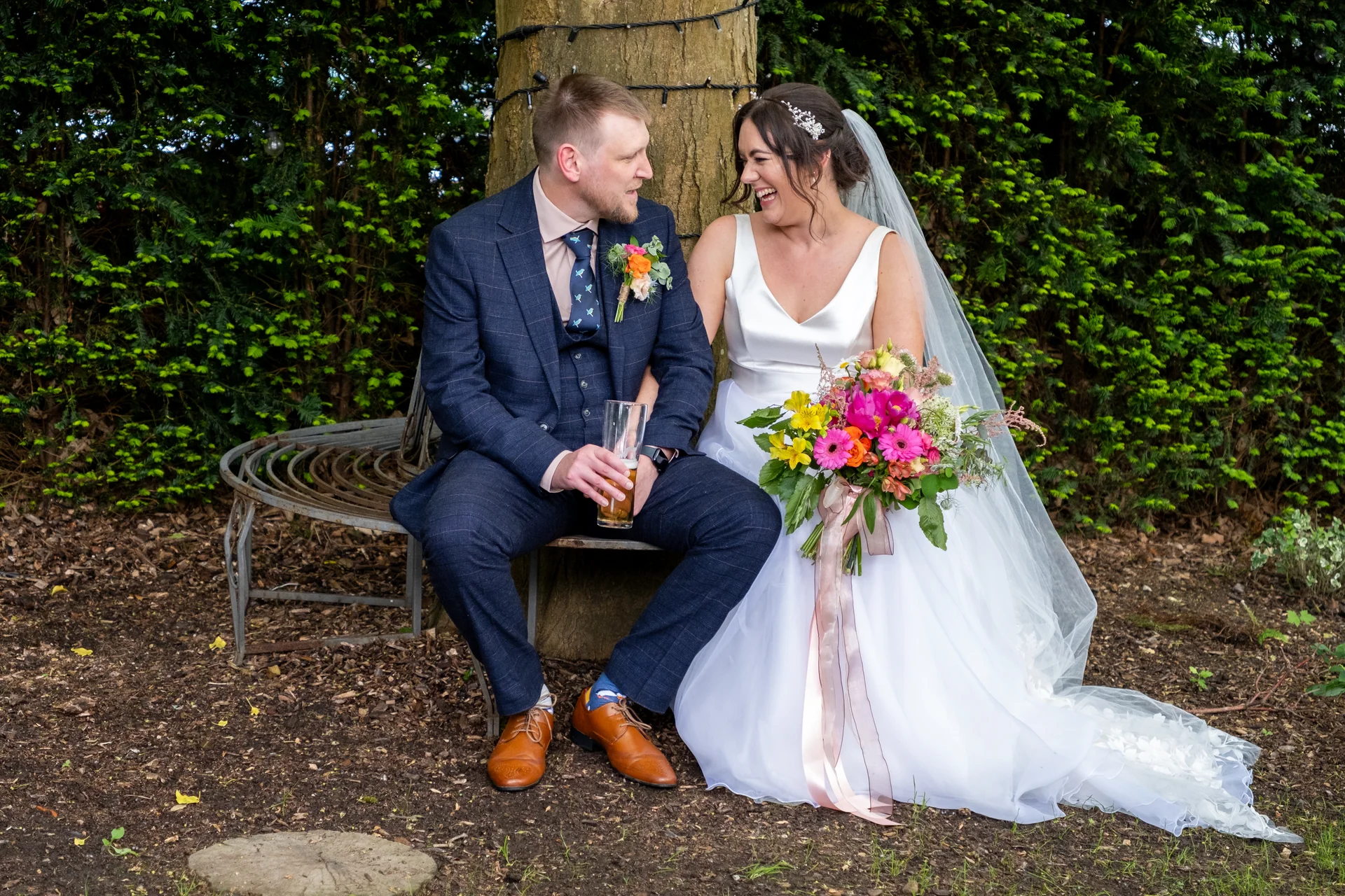 A newlywed couple shares a joyful laugh while sitting on a curved metal bench wrapped around a tree trunk, with the bride holding a vibrant colorful bouquet.