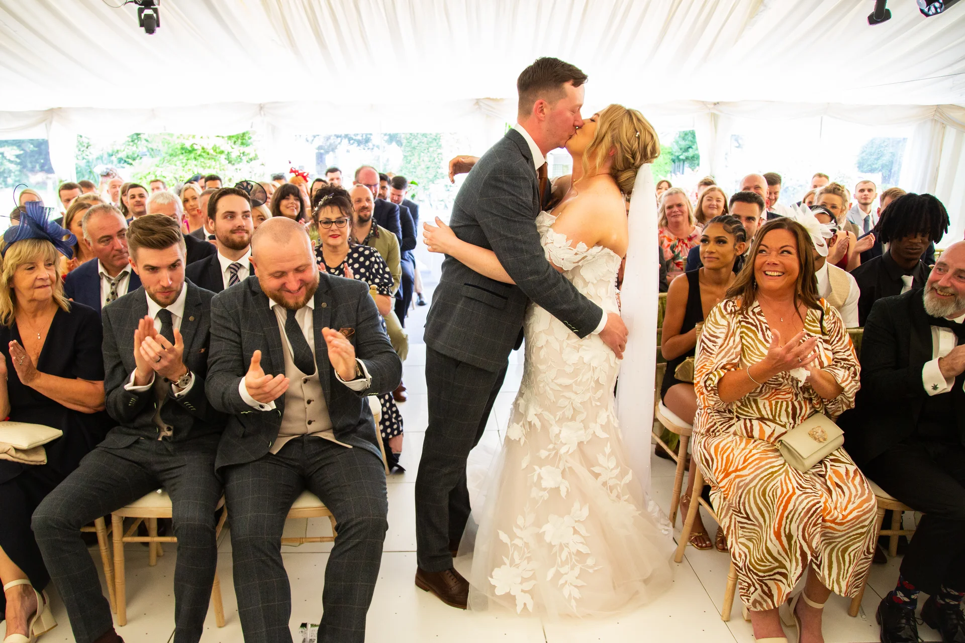 The newly married couple share their first kiss at the end of the aisle inside a bright marquee while seated guests clap and cheer with joy.