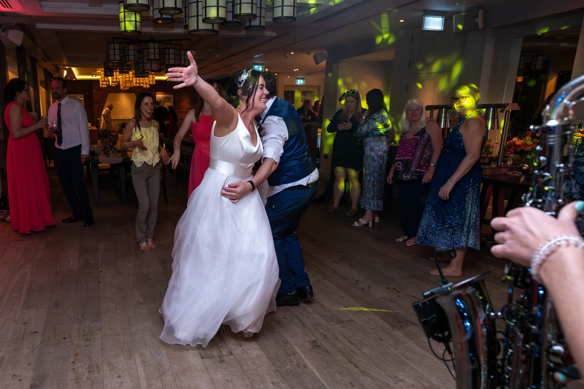 A joyful newlywed couple share their first dance on a wooden dance floor surrounded by wedding guests and lively disco lighting.