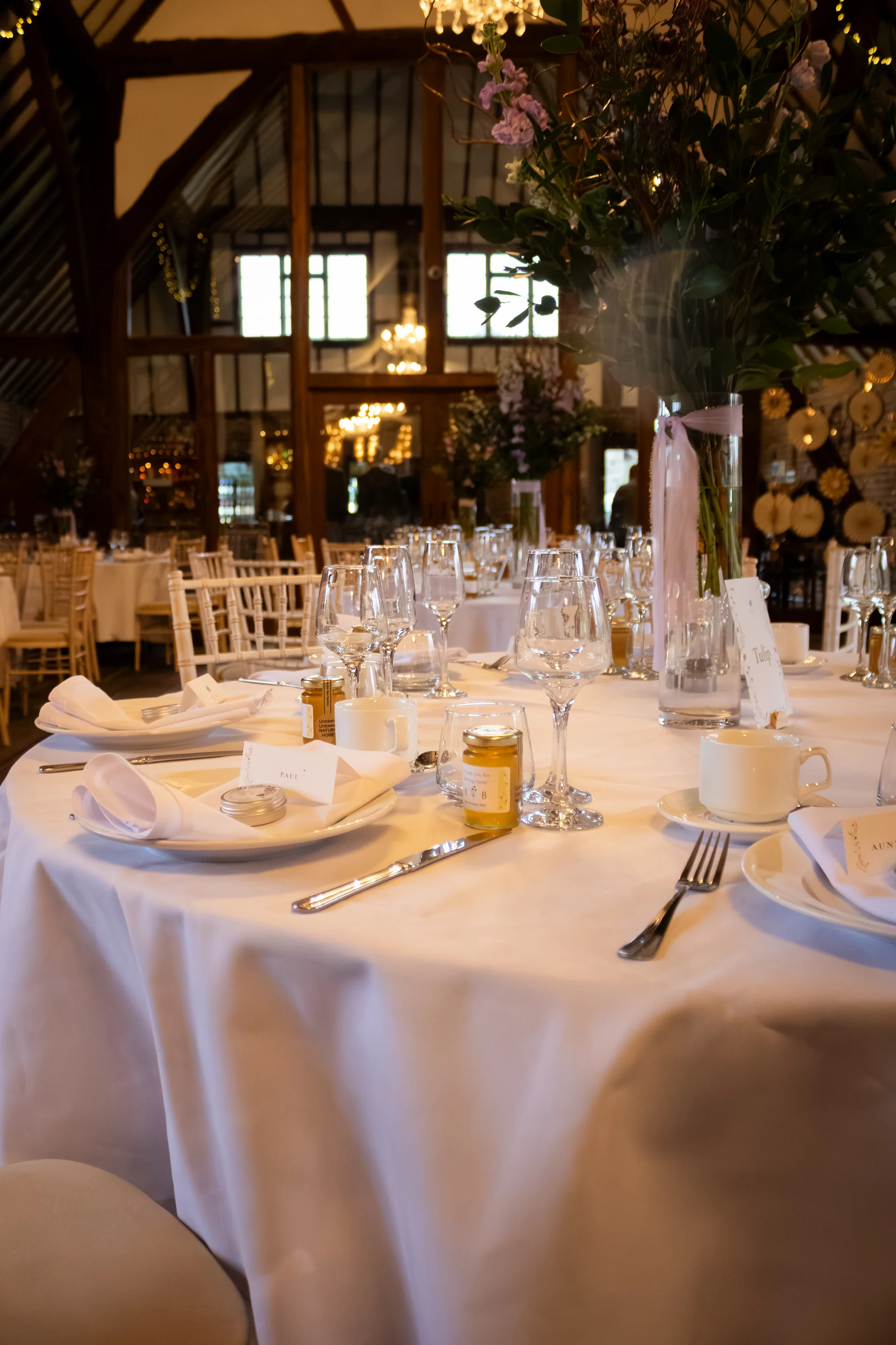 A beautifully set round wedding table inside a rustic oak-framed barn features crisp white linens, delicate glassware, place cards, and tall floral centerpieces.