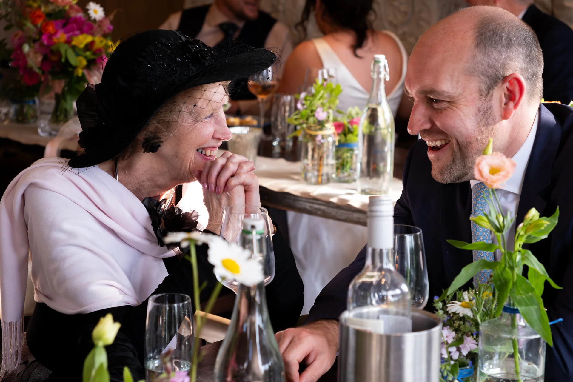A mature female guest wearing a black hat and fascinator shares a joyful, laughing moment with a male guest across a wedding reception table.