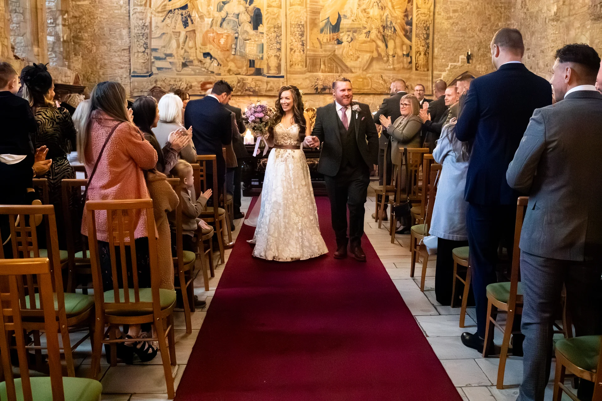 A newlywed couple walks hand in hand down a red aisle runner in a historic stone hall, smiling as guests applaud and celebrate around them.