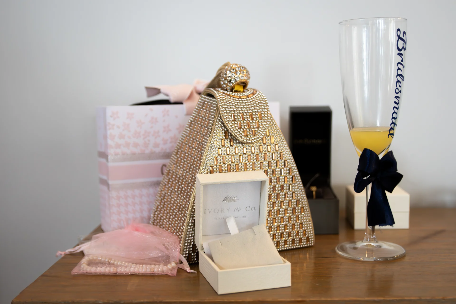 A thoughtful collection of bridal morning gifts, including a sparkly beaded clutch bag, an open Ivory and Co jewellery box, and a champagne flute bearing the word bridesmaid, rests on a wooden surface.