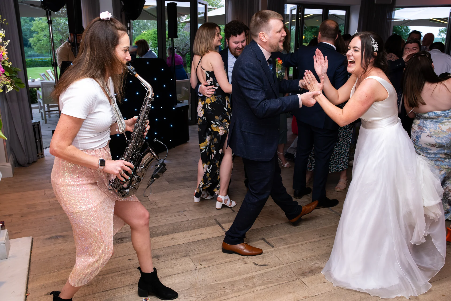 A joyous bride and groom laugh and clap as they dance on the wooden floor, accompanied by a live saxophonist playing nearby at an evening wedding reception.
