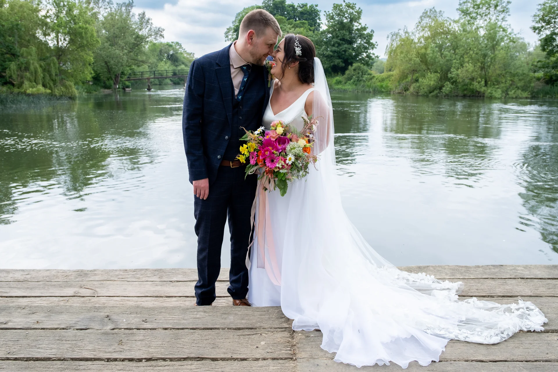 A newlywed couple share a tender nose-to-nose moment while standing on a wooden jetty by a tranquil lake, with the bride holding a vibrant wildflower bouquet and wearing a flowing white gown and long veil.